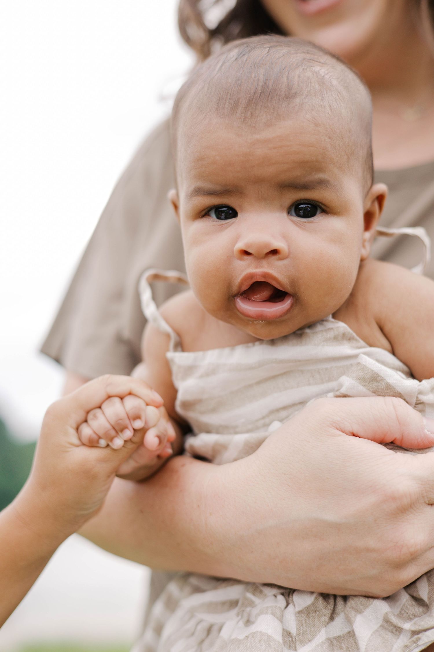 A mother in a beige dress holds her wide-eyed infant baby, who grips her finger outdoors.