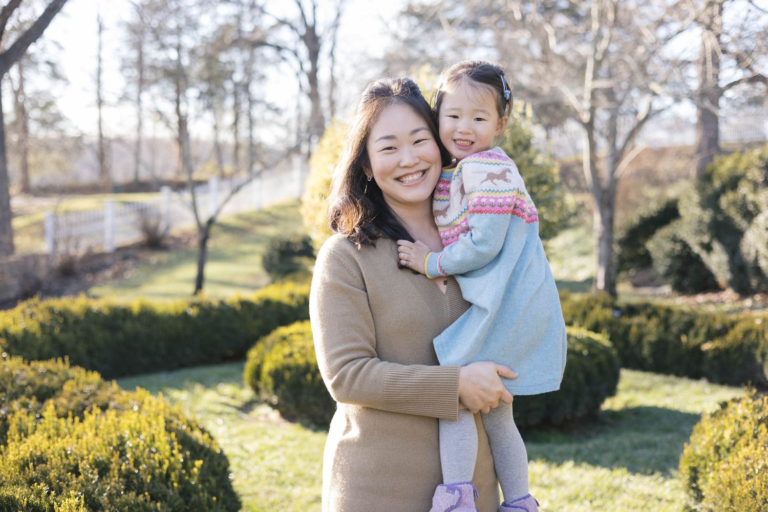 A parent and child share a joyful moment together in a sunny garden with manicured shrubs and trees in the background.