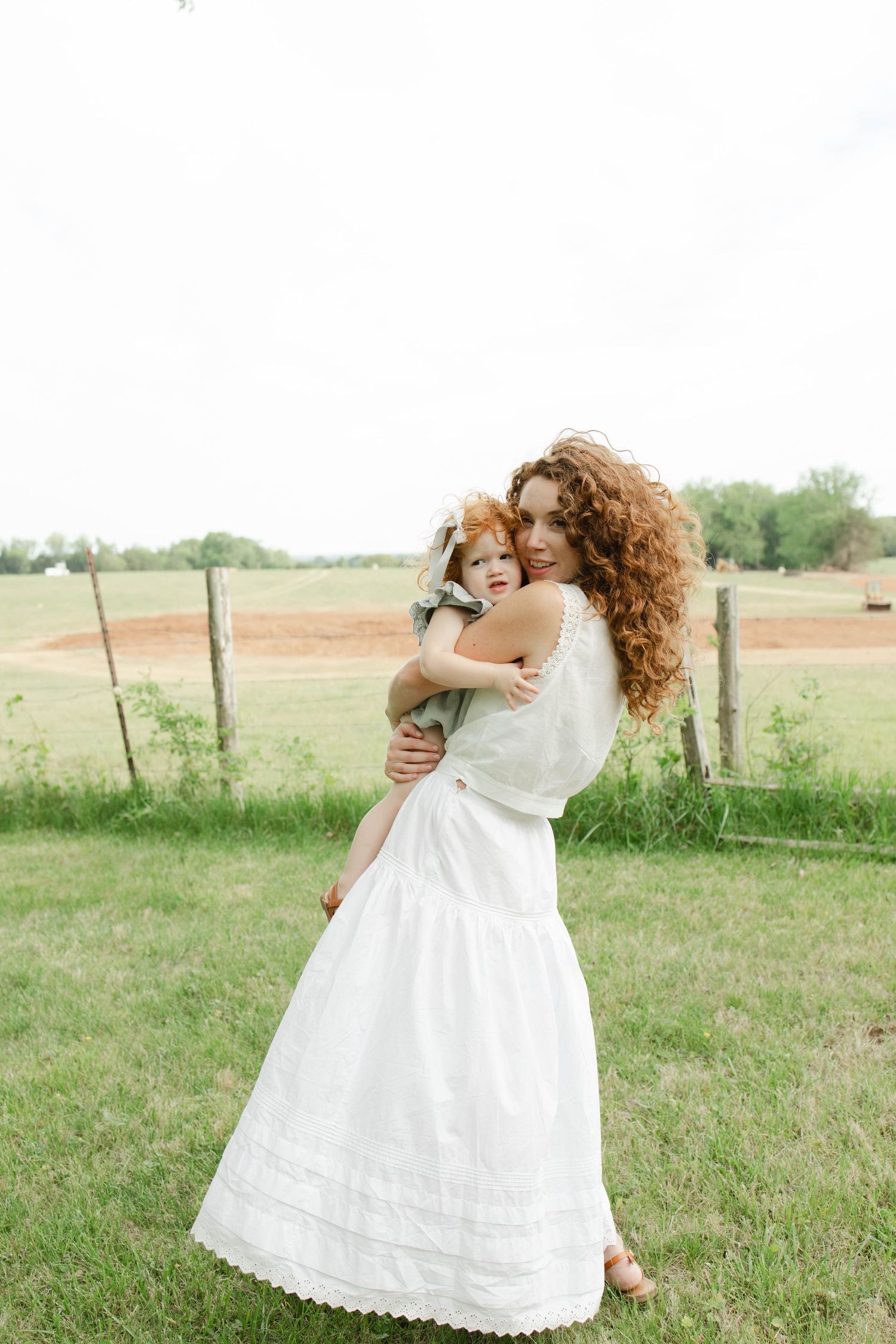 A curly-haired woman in a white dress holds a young child in a green farm field.