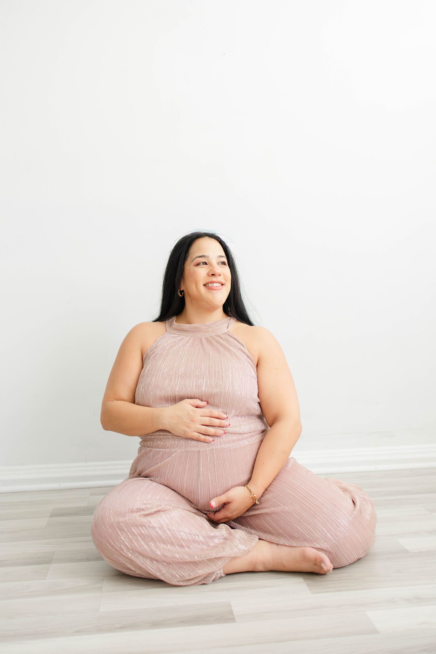 Woman in pink dress sits cross-legged on floor during maternity session.