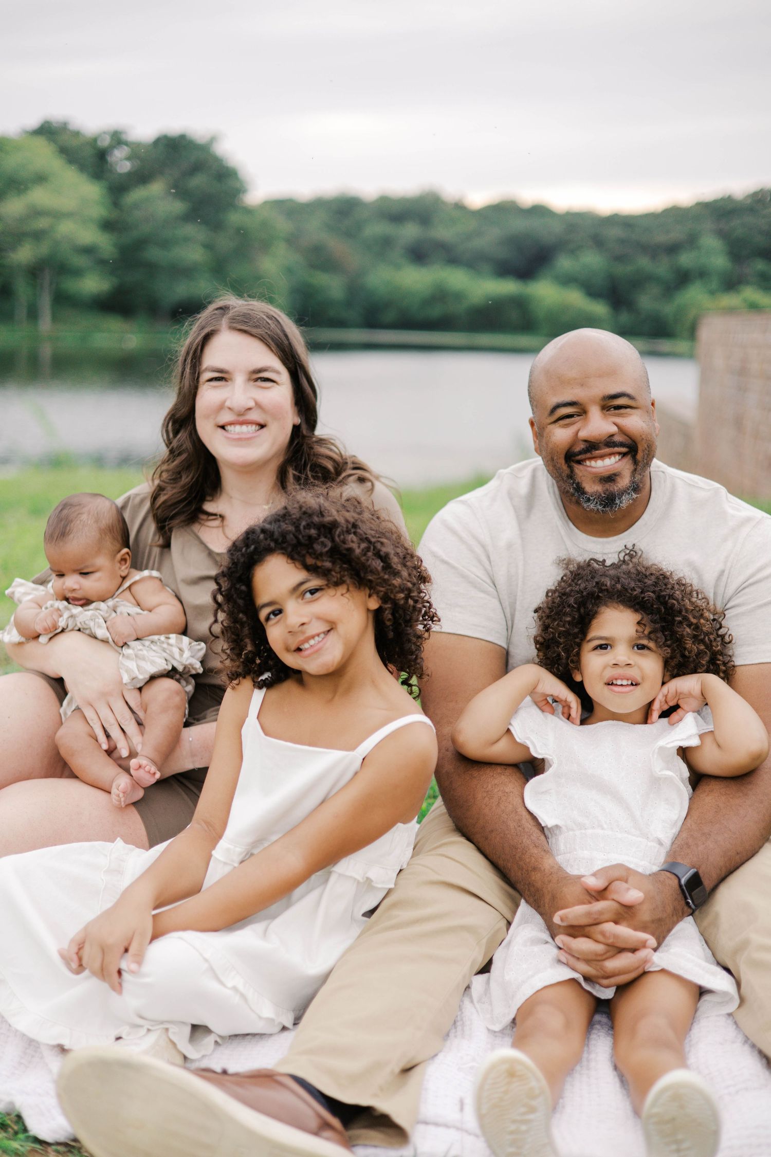 Happy interracial family of five posing outdoors near a lake, with two curly-haired girls and a newborn baby.