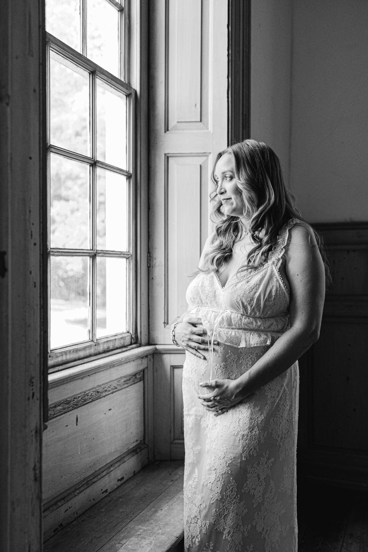 Black and white maternity portrait of a pregnant woman in a lace gown cradling her belly beside a sunlit window.