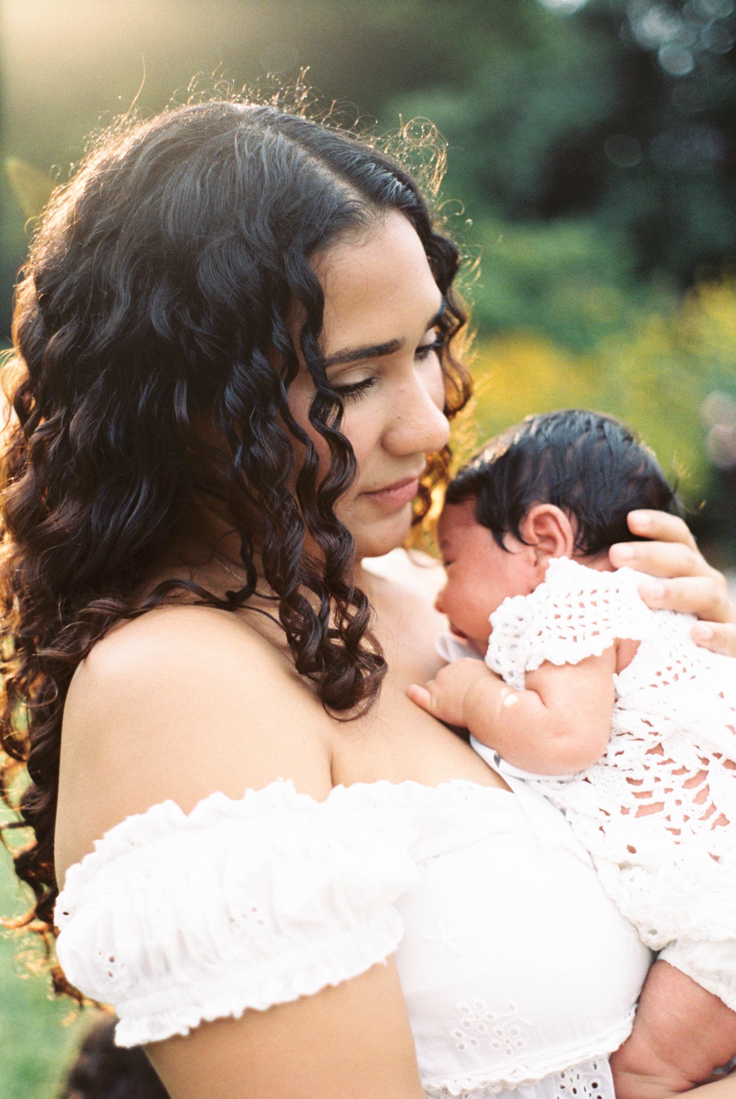 A young mother with curly dark hair in a white off-shoulder dress tenderly holds her newborn baby outdoors in golden light.