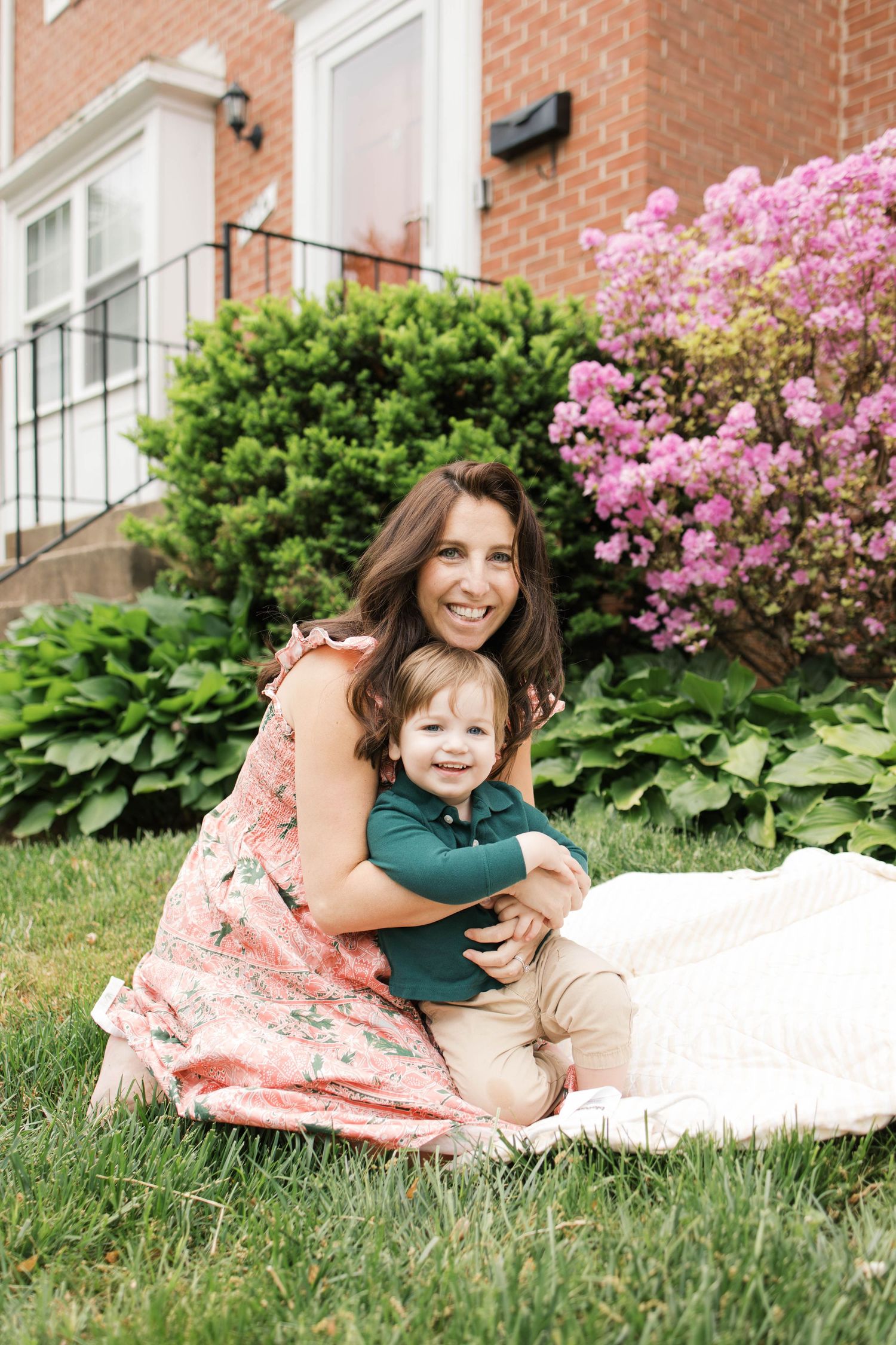 A smiling woman in a floral dress hugs a toddler boy on a blanket in a lush garden beside a brick home.