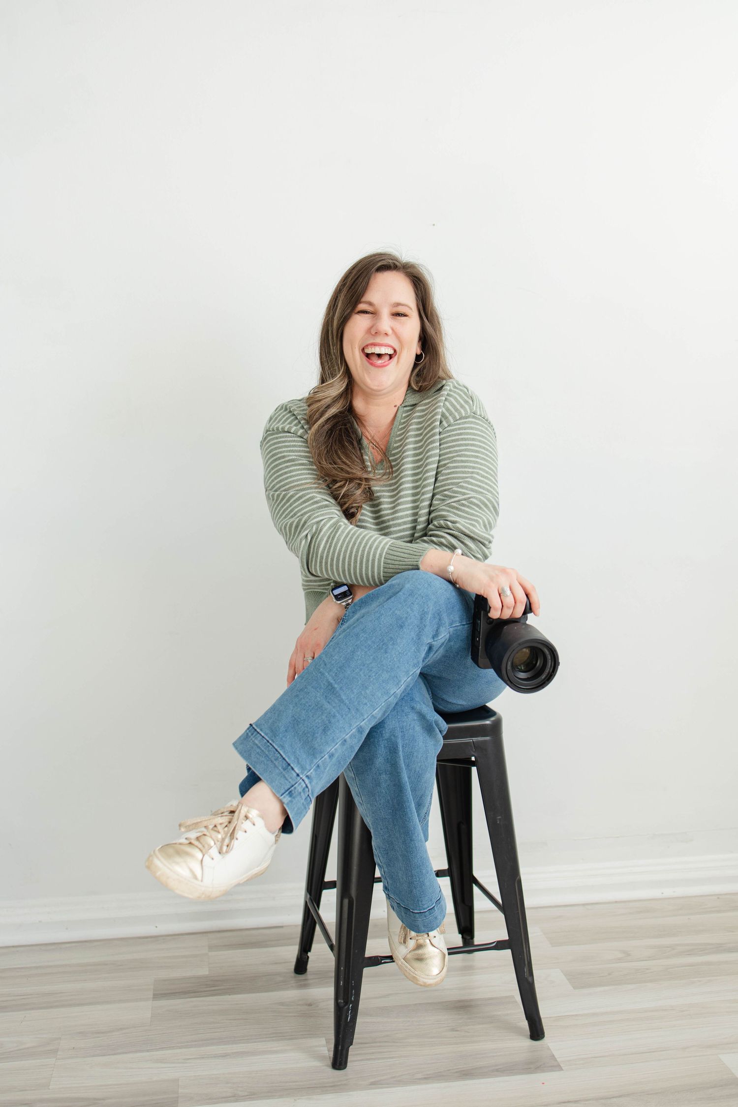 Woman in casual attire sits on black stool holding camera in bright studio setting.