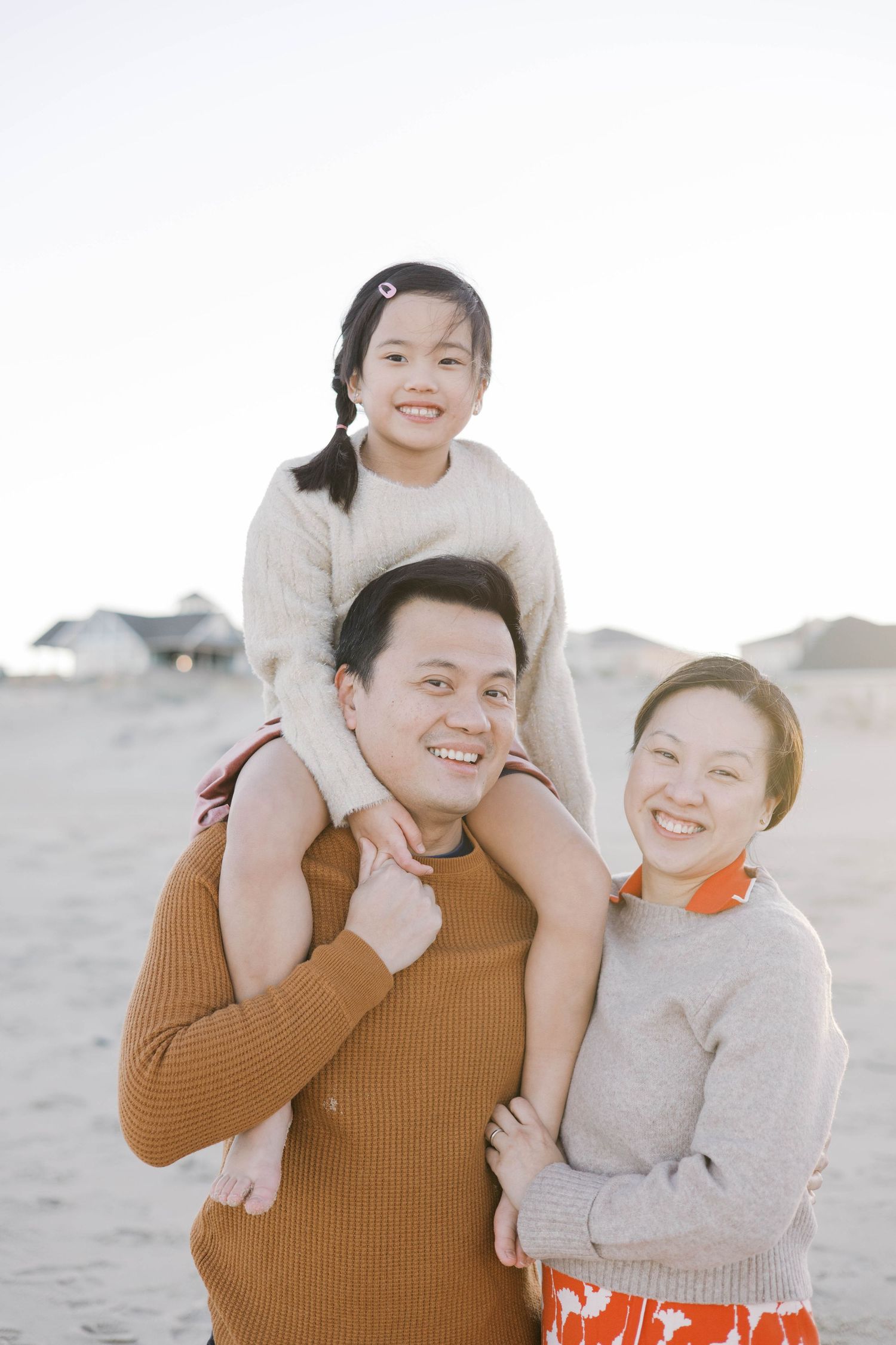 Happy Asian family of three posing on a sandy beach, with a young girl sitting on her father's shoulders while mother smiles beside them.
