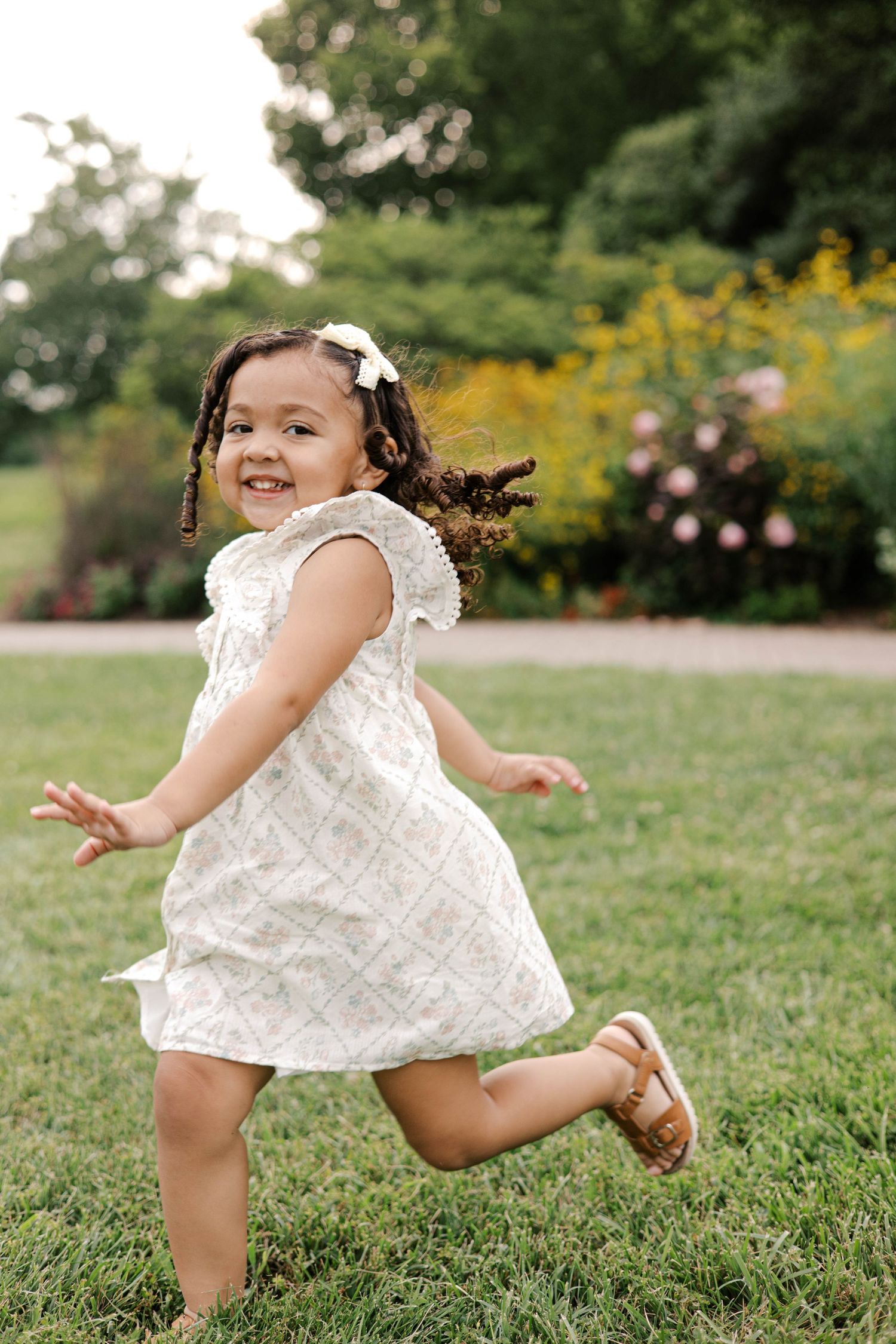 A joyful toddler girl in a white dress runs through a lush green park with colorful flowers in the background.