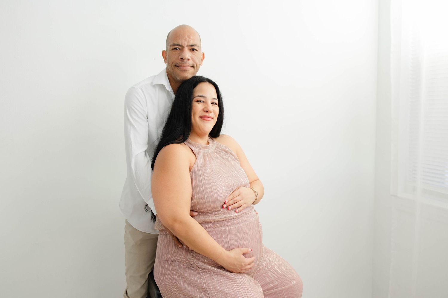 Couple embraces during maternity photoshoot against white backdrop.