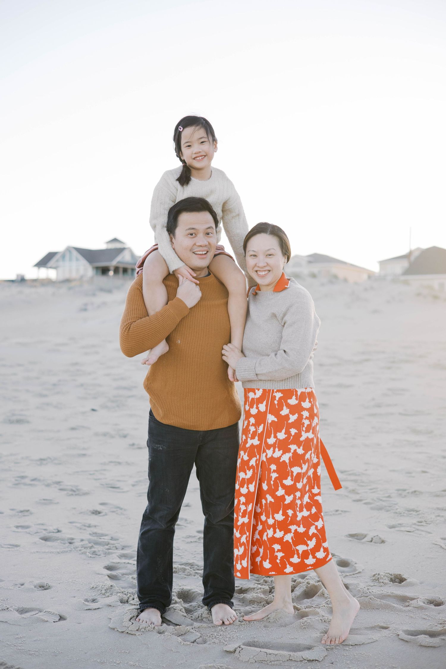 Family enjoying a playful moment together on a sandy beach with houses in background.