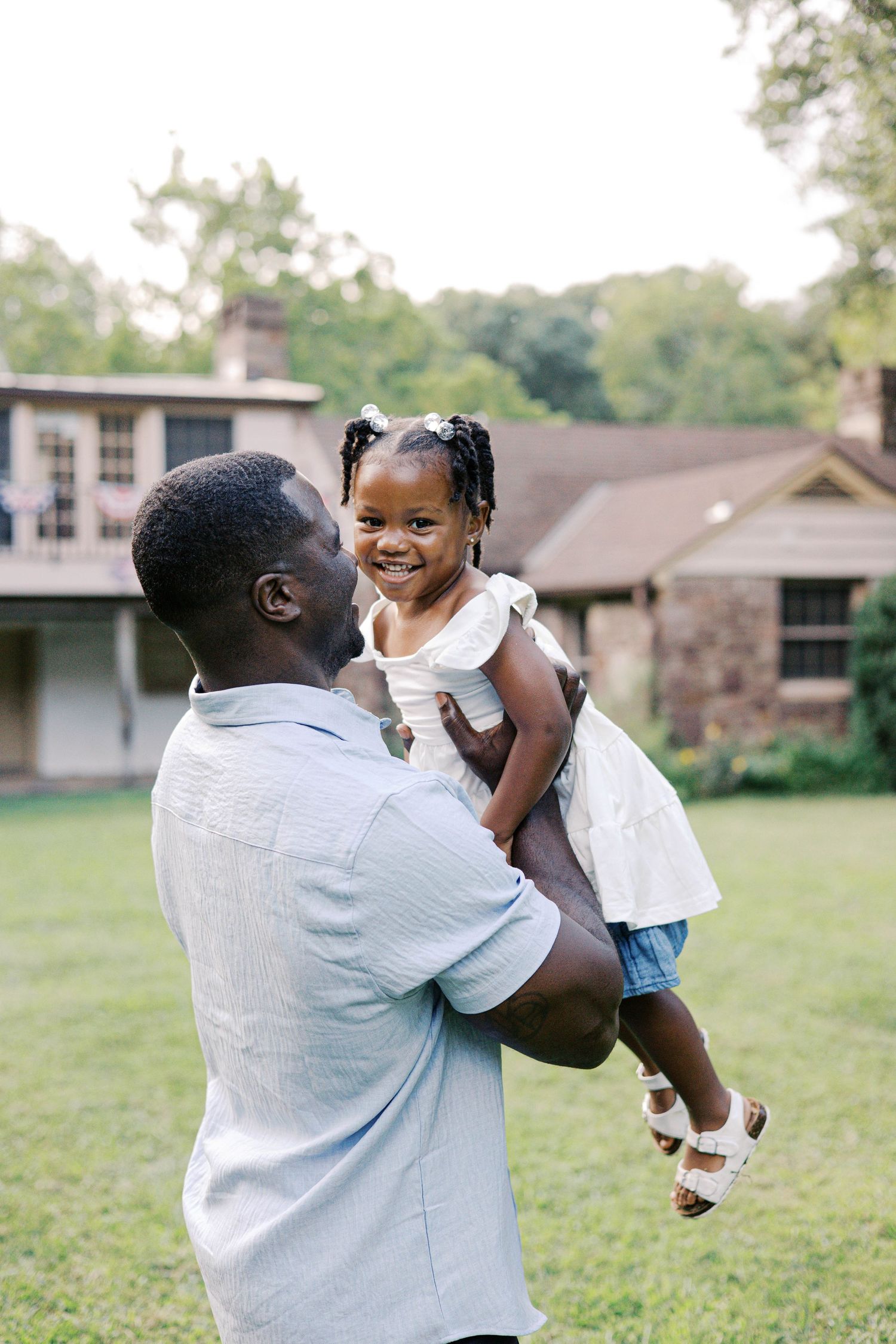 A joyful Black father lifts his smiling little girl in a white dress outdoors near a brick house.