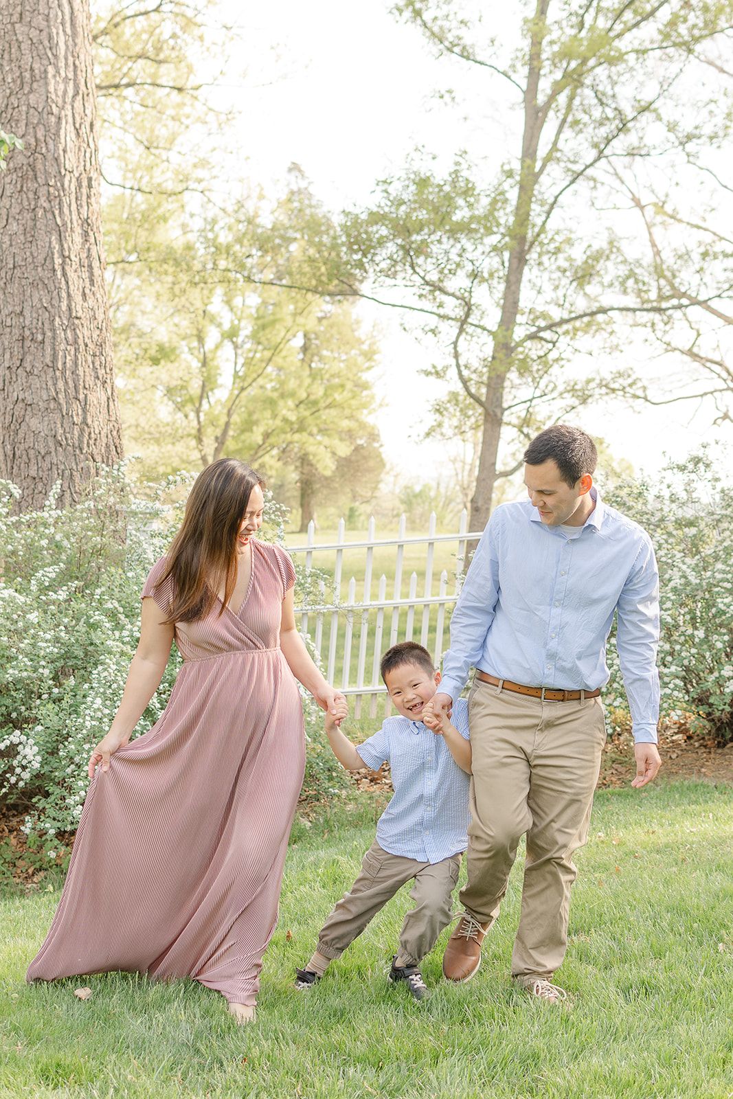 A family of three walks hand-in-hand on green grass, mom in mauve gown, dad in blue shirt, swinging their young son.