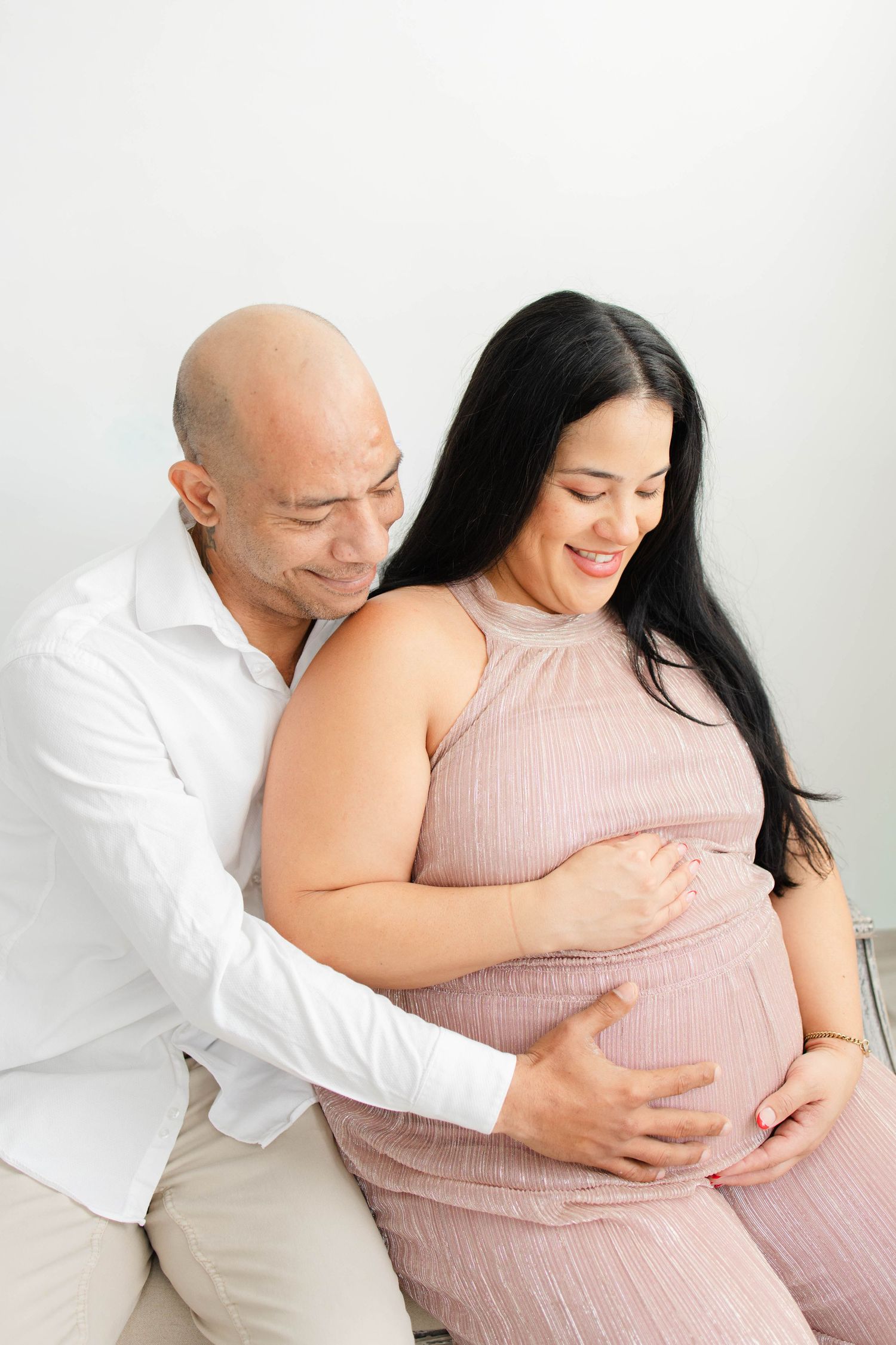 Couple embraces in a tender maternity photo shoot against white backdrop wearing pink dress and white shirt.