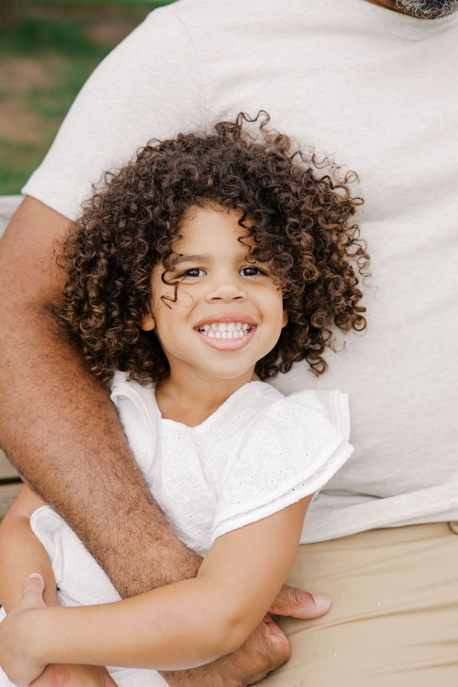 A young girl with curly hair smiles brightly while being held by an adult in a white shirt outdoors.
