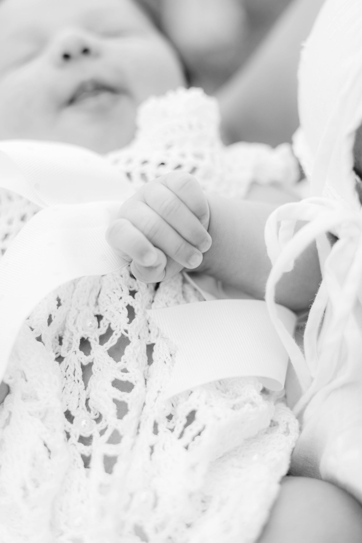 Black and white photo of a newborn baby's tiny hand clutching lace fabric of a white christening gown.