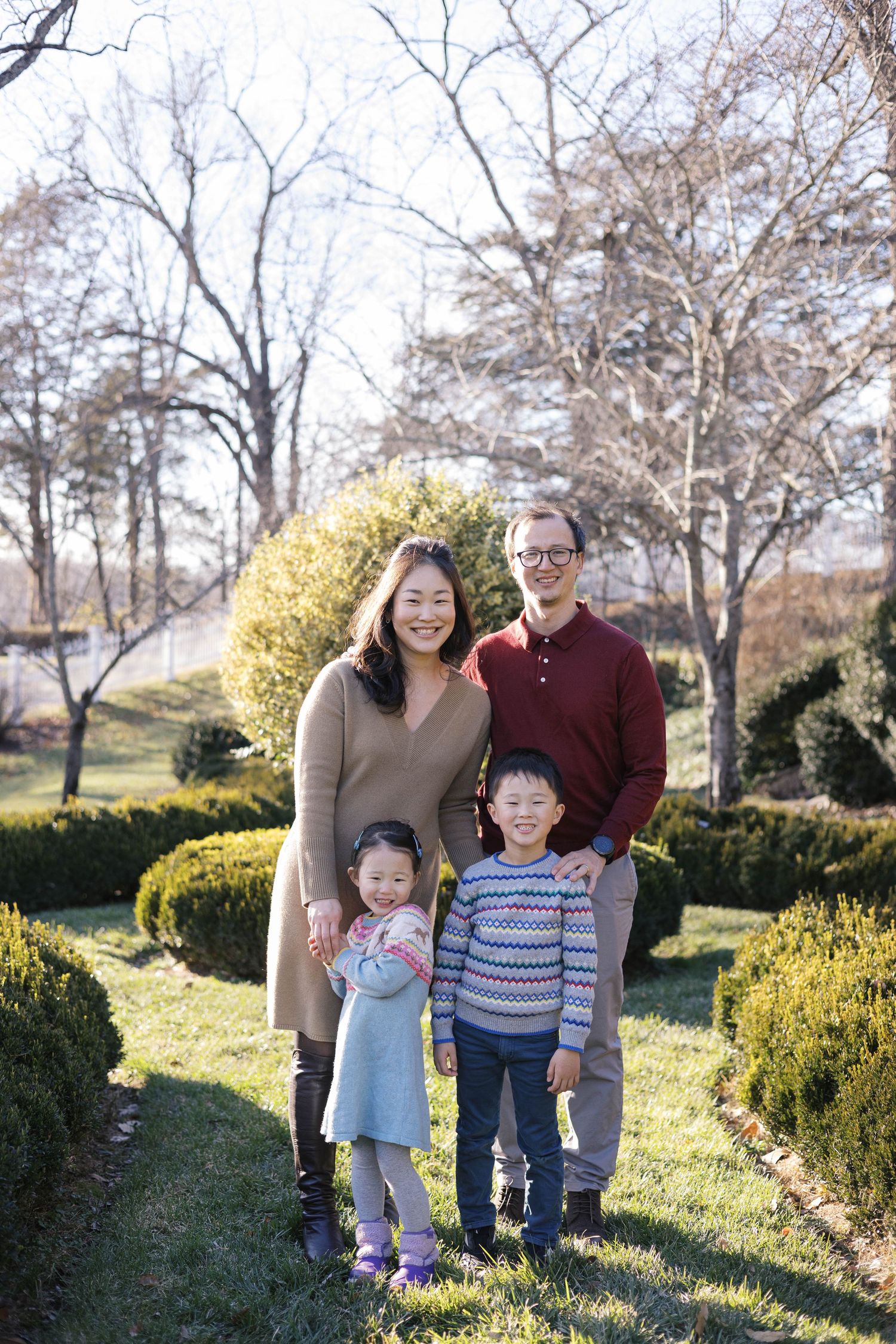 Family portrait in a garden setting with evergreen shrubs and bare winter trees.