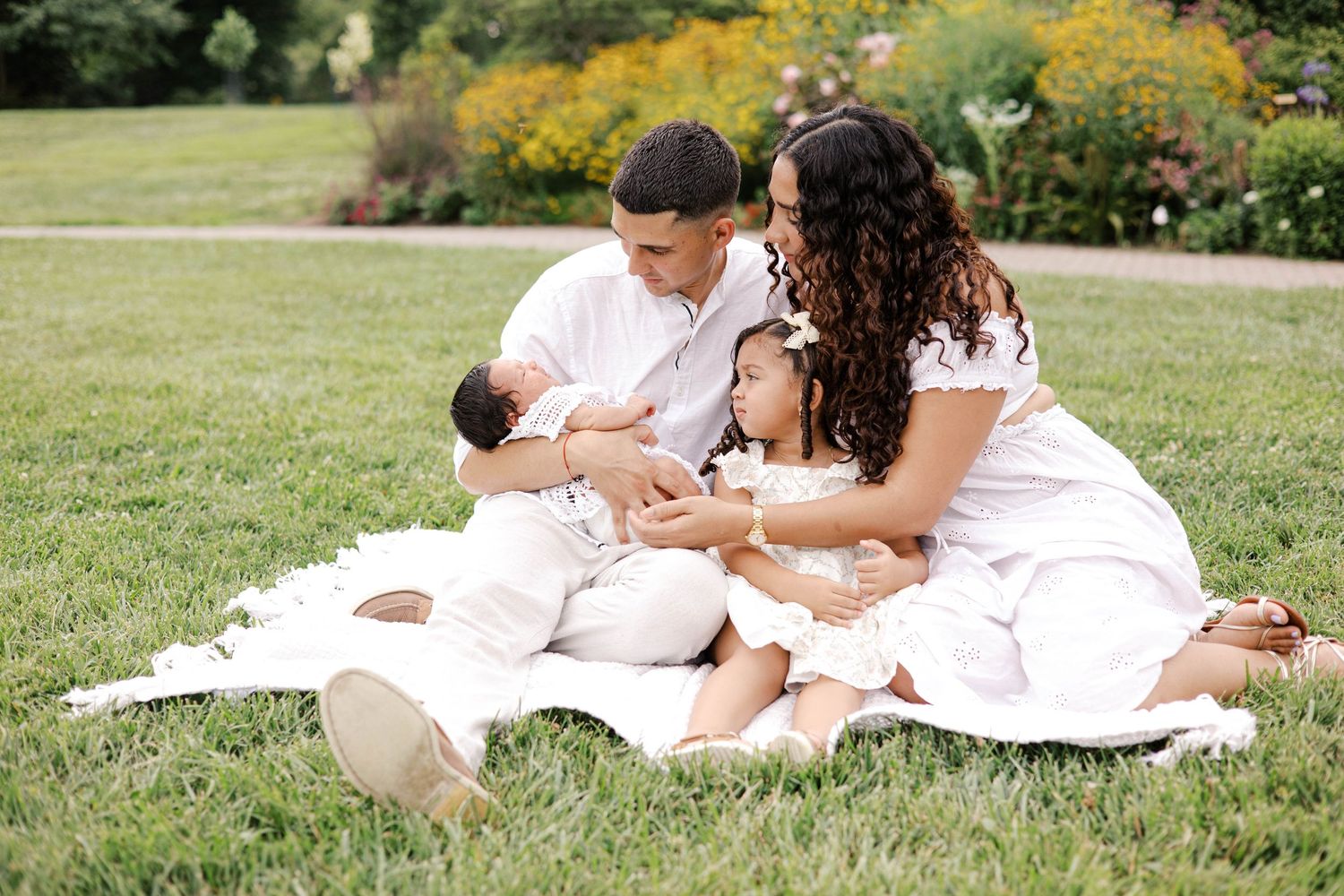 Family of four in white outfits sitting on blanket in lush green park, parents lovingly holding baby and toddler.