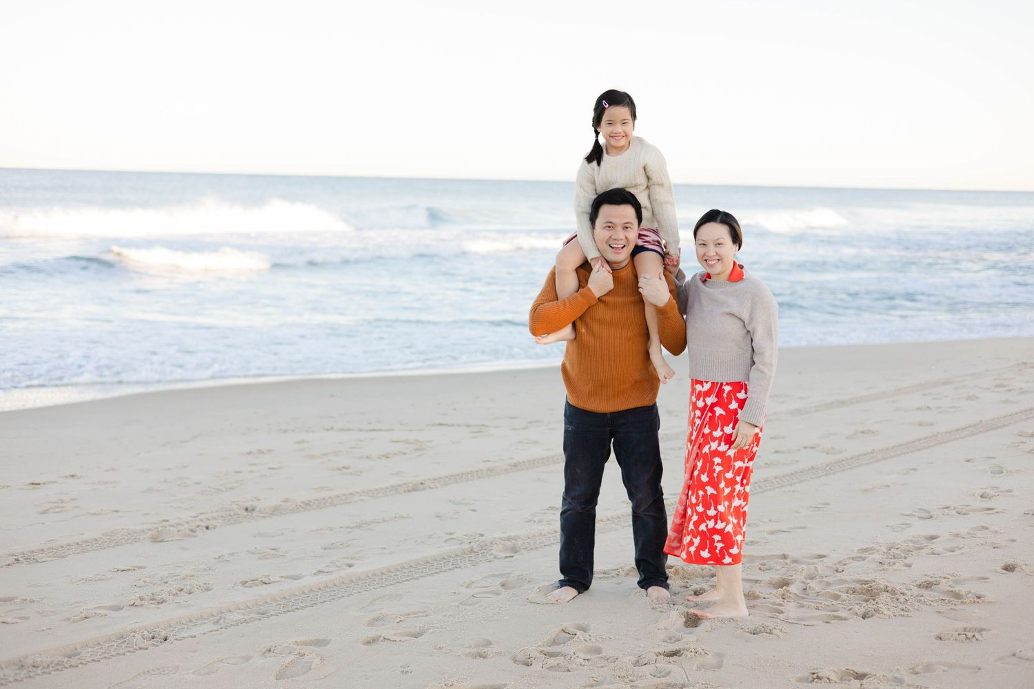 Happy Asian family of three enjoying a sunny beach day, with daughter sitting on father's shoulders near the ocean waves.