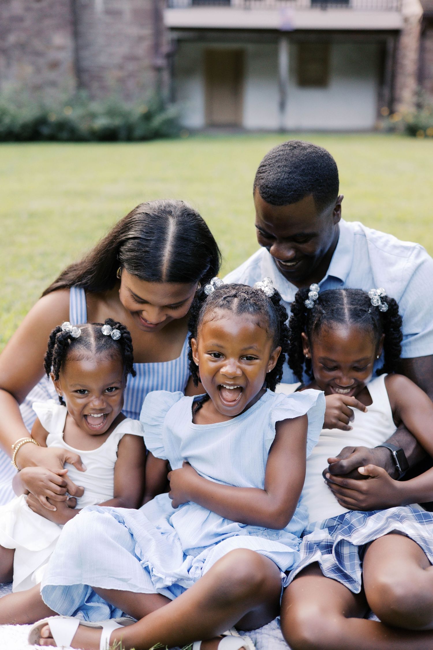A joyful Black family with two parents and three young daughters in matching blue outfits sitting together on grass outdoors.