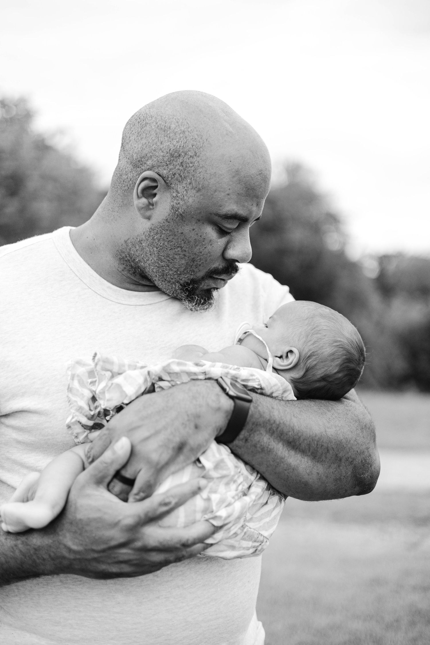 Black and white photo of a bald bearded man tenderly cradling a newborn baby outdoors.