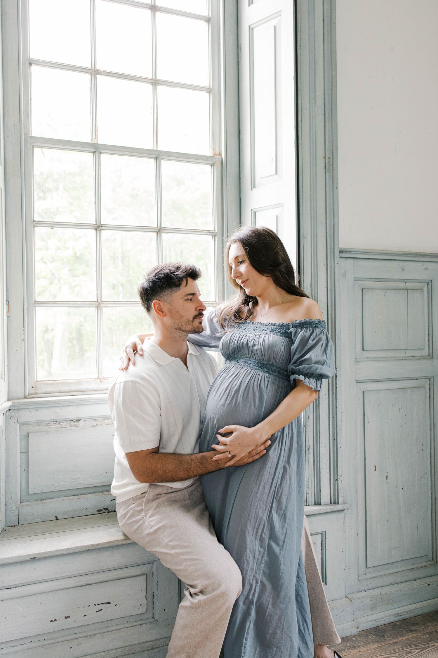 Couple posing for maternity photo near window, woman in blue off-shoulder dress cradling baby bump, man in white outfit.