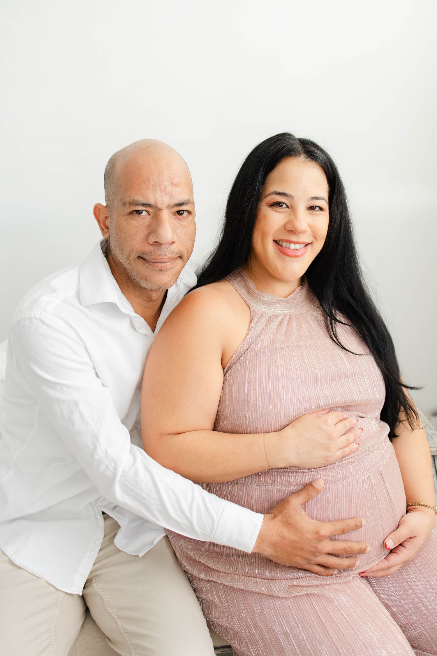 Expectant couple poses together against white wall in coordinated outfits.