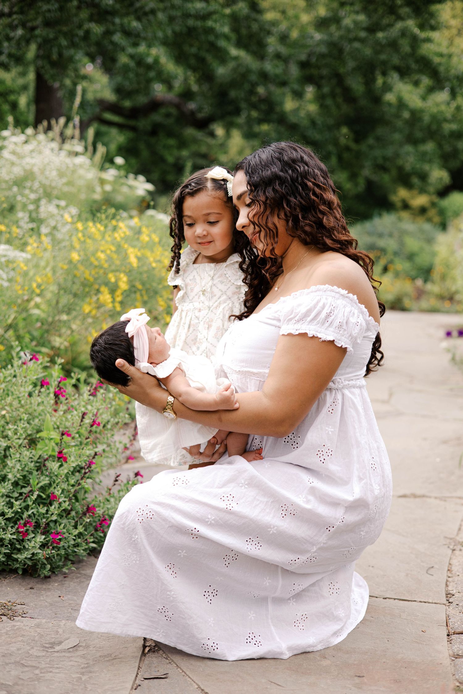 A mother in a white dress sits in a garden holding a newborn while her young daughter in white looks on lovingly.