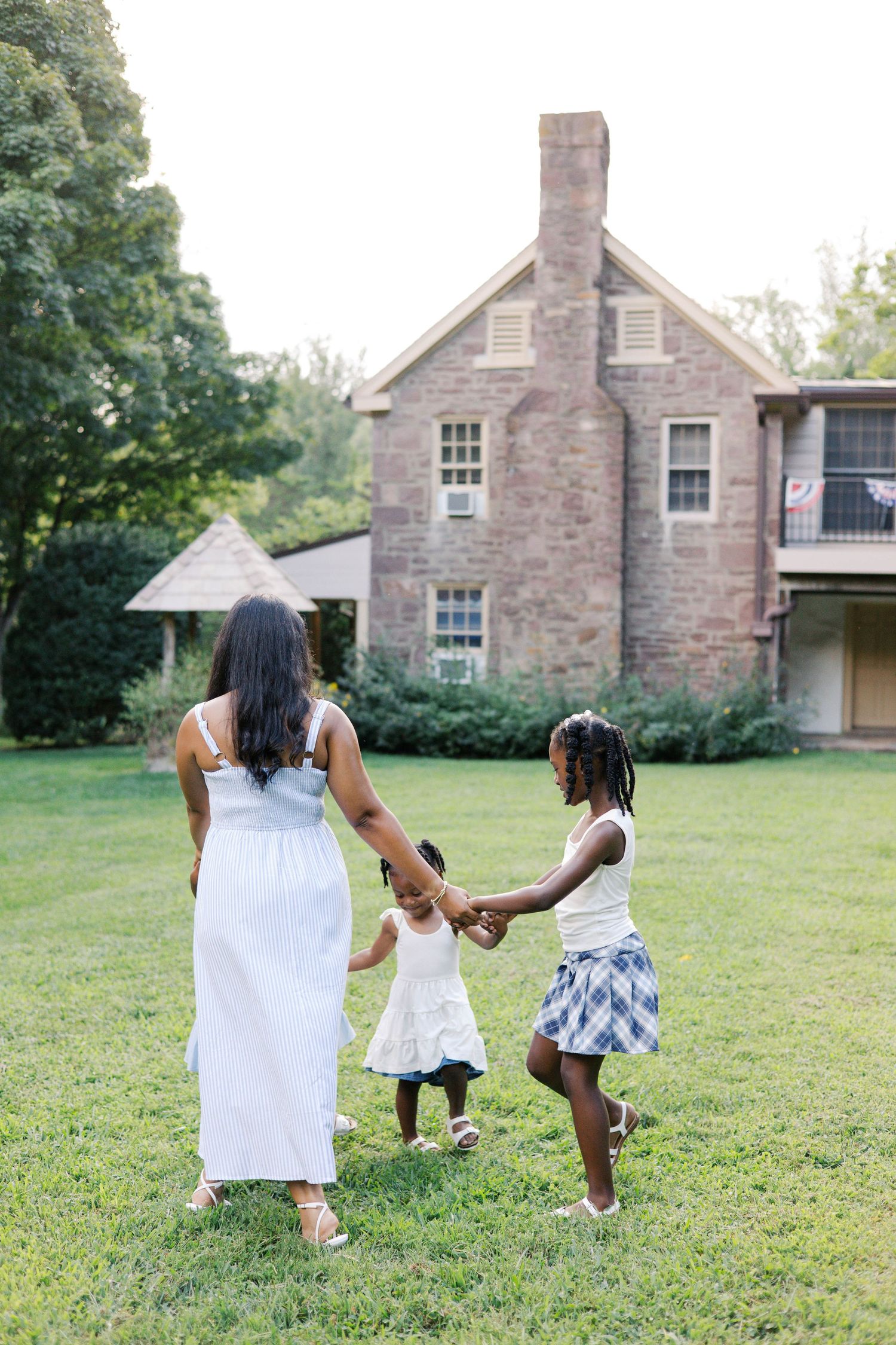A mother and two daughters walk hand-in-hand across a green lawn toward a historic brick house.