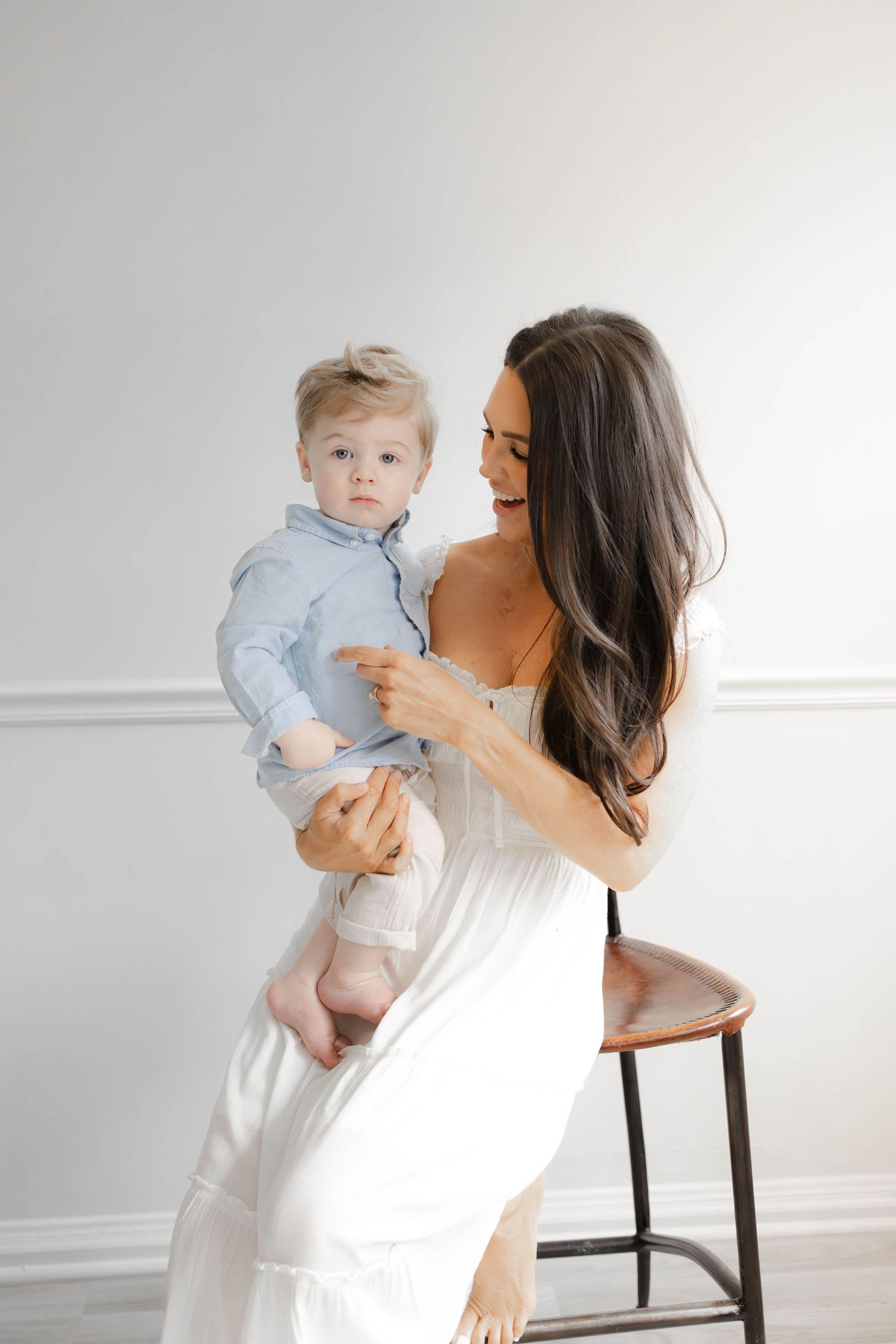 A smiling brunette woman in a white dress holds a baby boy in a blue shirt while sitting on a stool.
