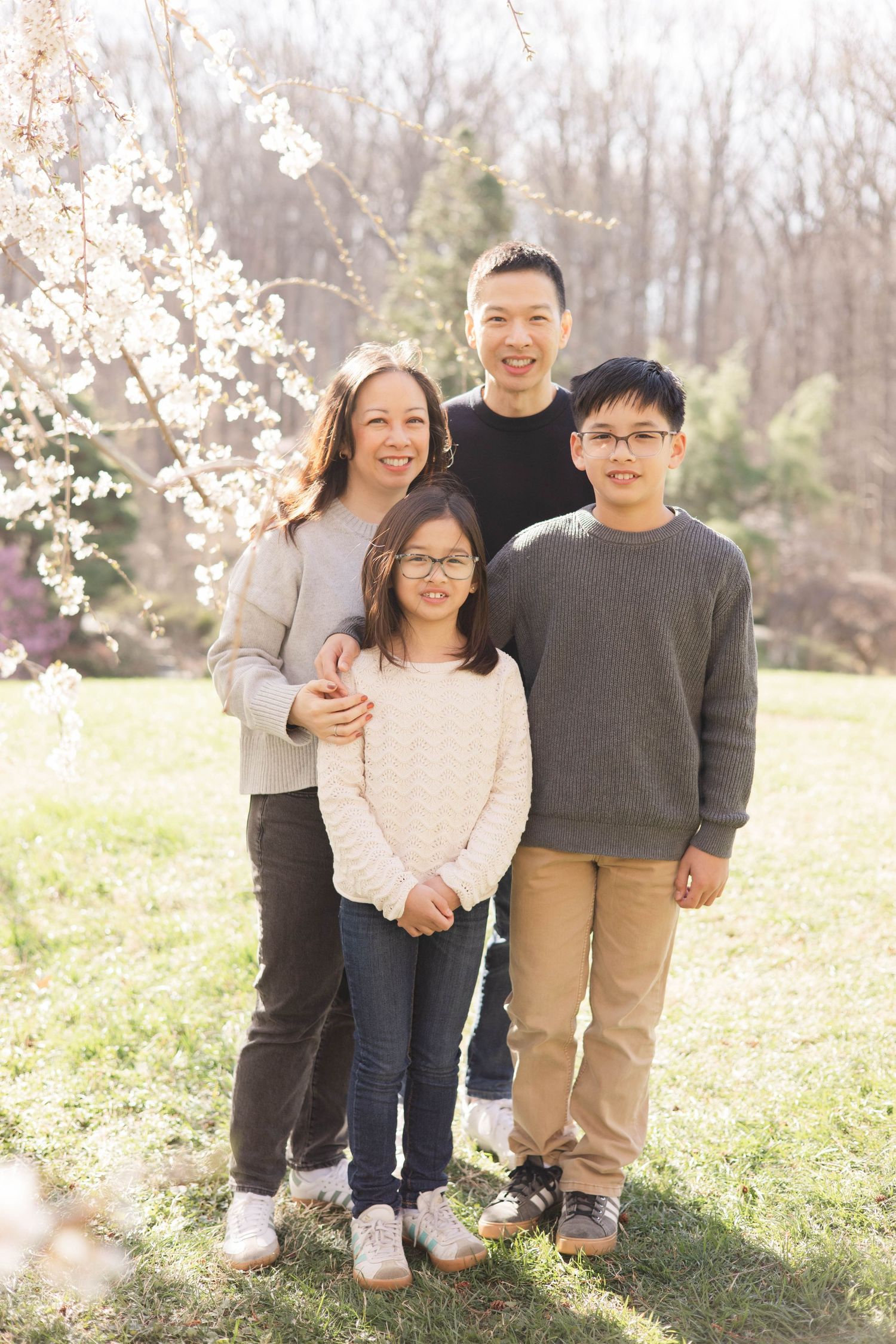 Family of four smiling outdoors near blooming cherry tree in spring, parents with two children wearing glasses.