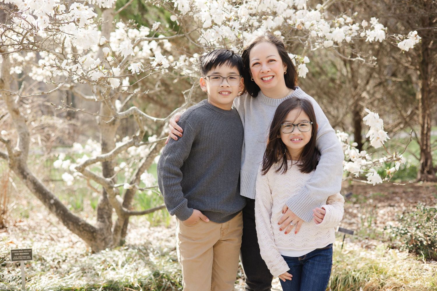 A smiling Asian family of three poses under blooming white cherry blossom trees in a spring garden.