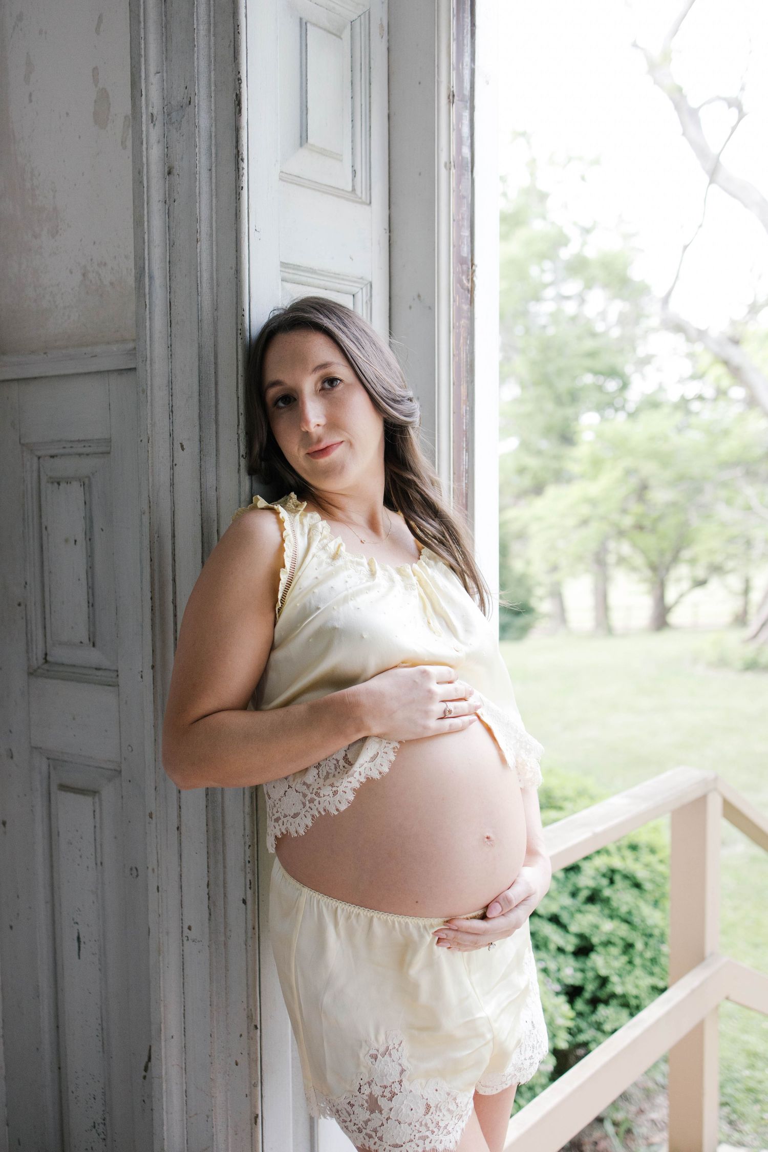 Pregnant woman in yellow dress cradling baby bump, leaning against rustic door frame on a porch.