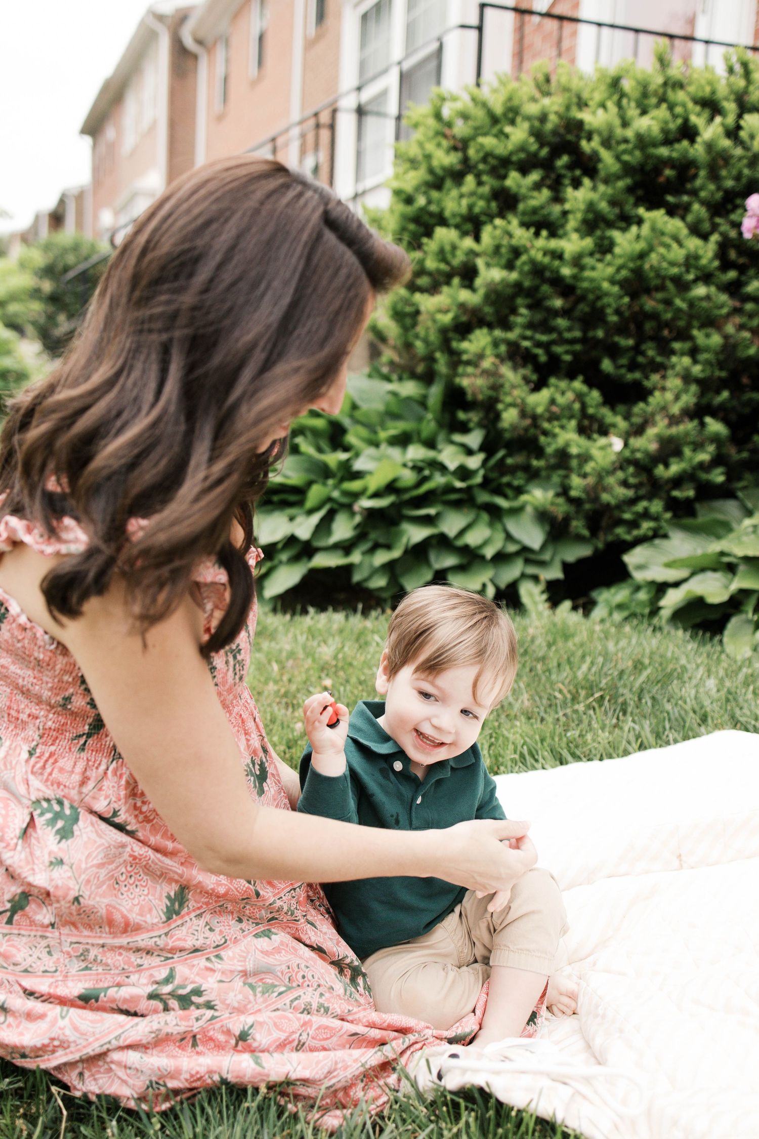 A mother in a floral dress sits on a blanket outdoors with her smiling toddler in a green shirt.