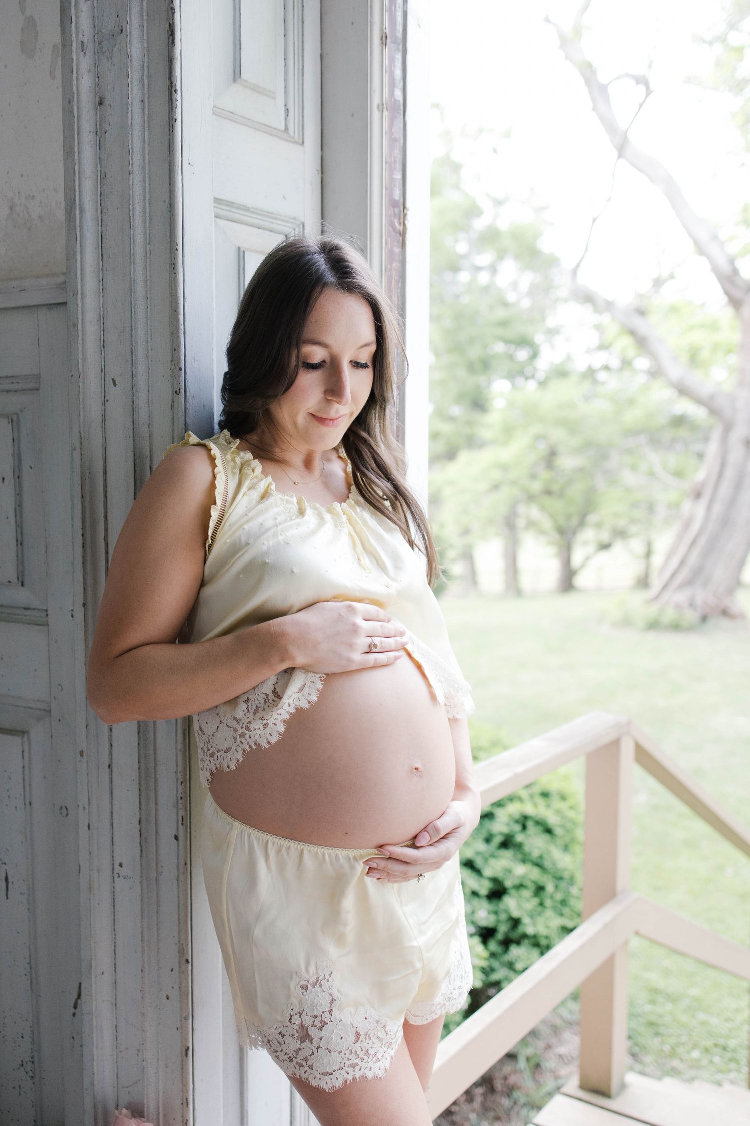 Pregnant woman in yellow lace outfit cradling her baby bump by a vintage door with green trees in background.