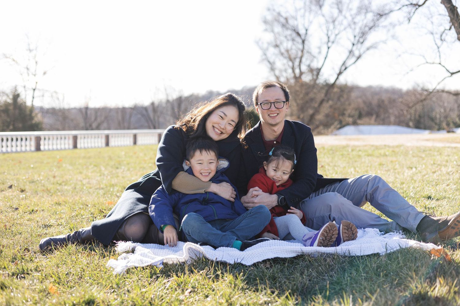 Family of four smiling and sitting together on a blanket in a grassy park on a sunny autumn day.