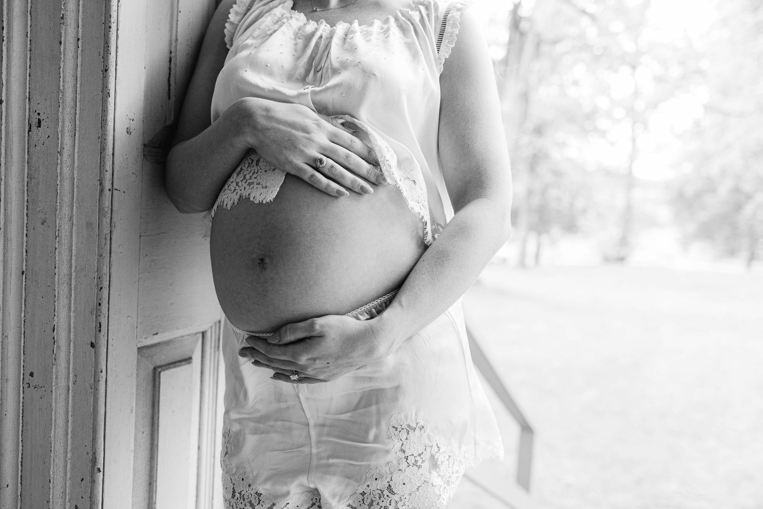 Black and white maternity photo of pregnant woman cradling baby bump in delicate lace dress.