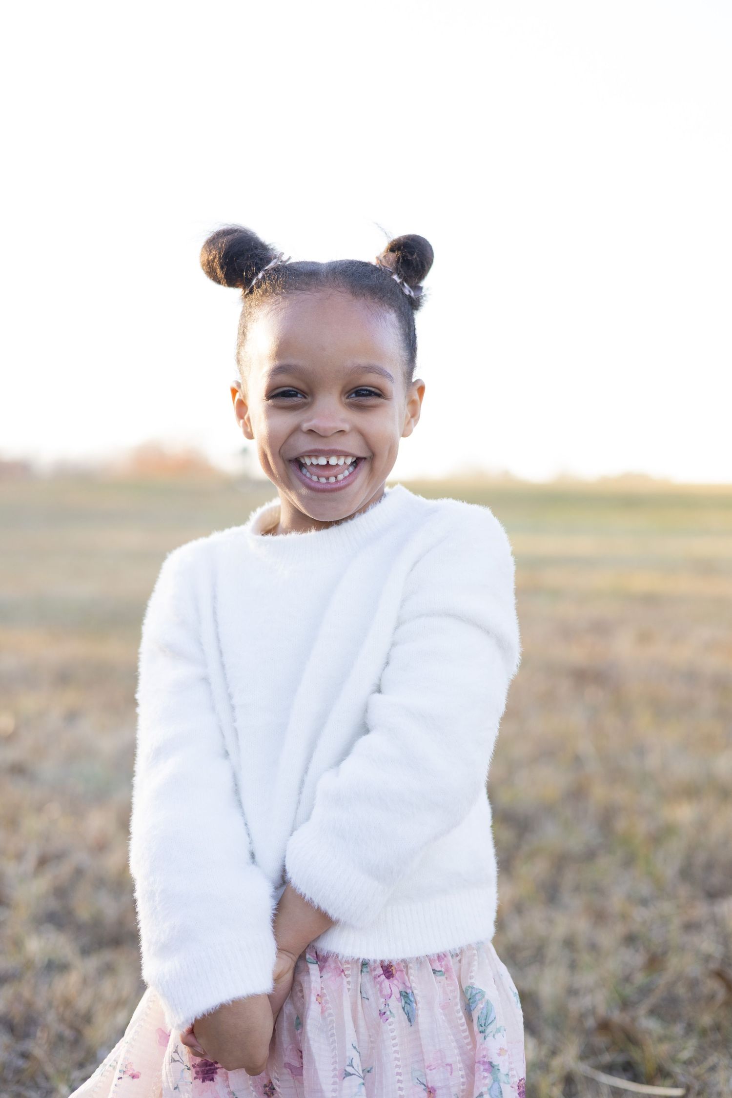 Young child in white sweater poses playfully in a grassy field at sunset.