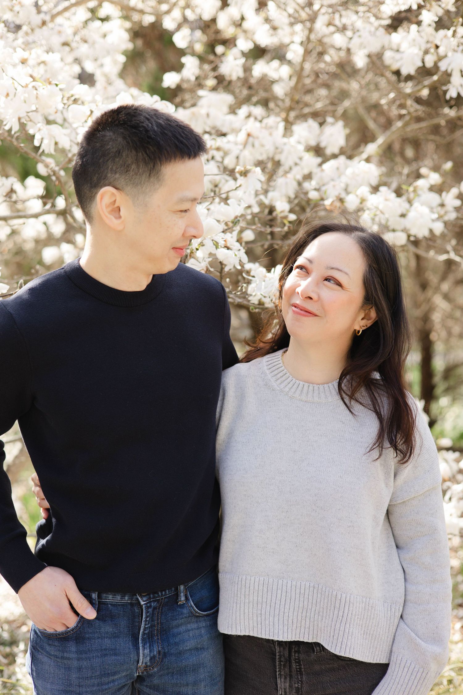 A couple smiles at each other under blooming white cherry blossom trees in spring.