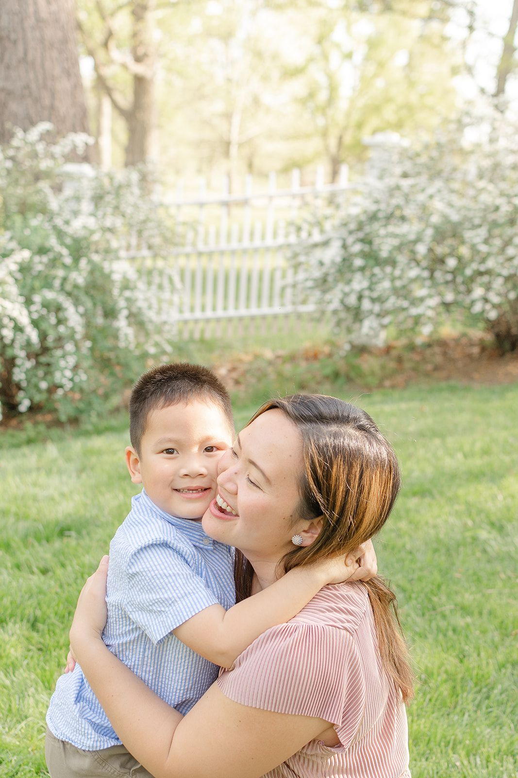 A mother hugs and kisses her smiling young son outdoors on green grass near a white picket fence.