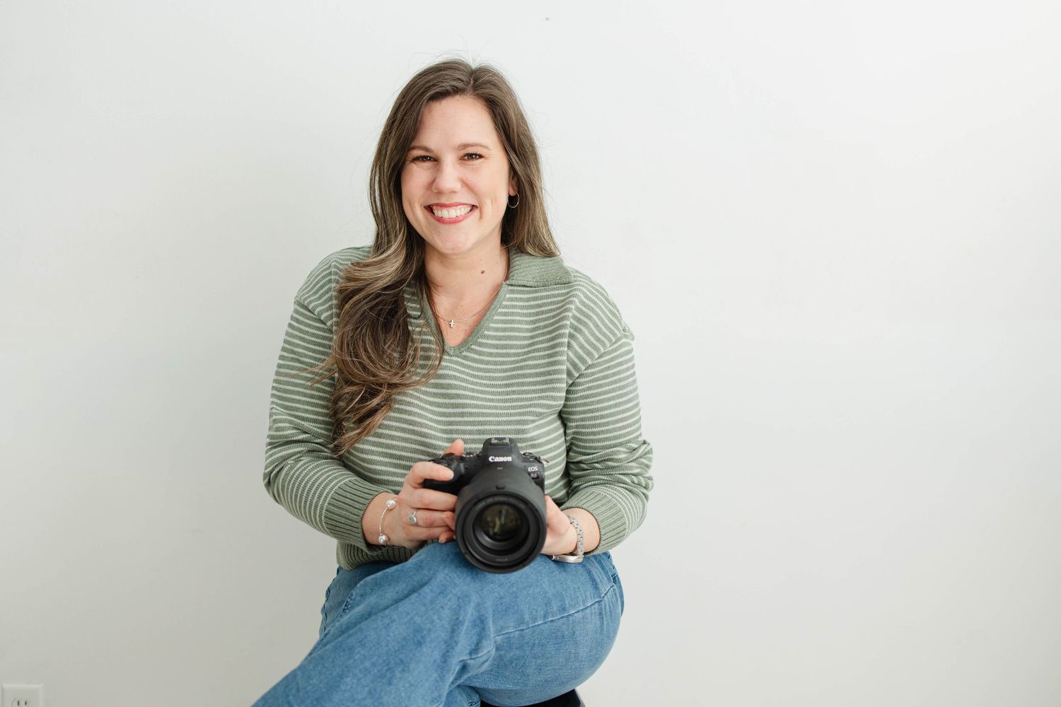 Photographer in green striped shirt poses with professional camera equipment in bright studio setting.
