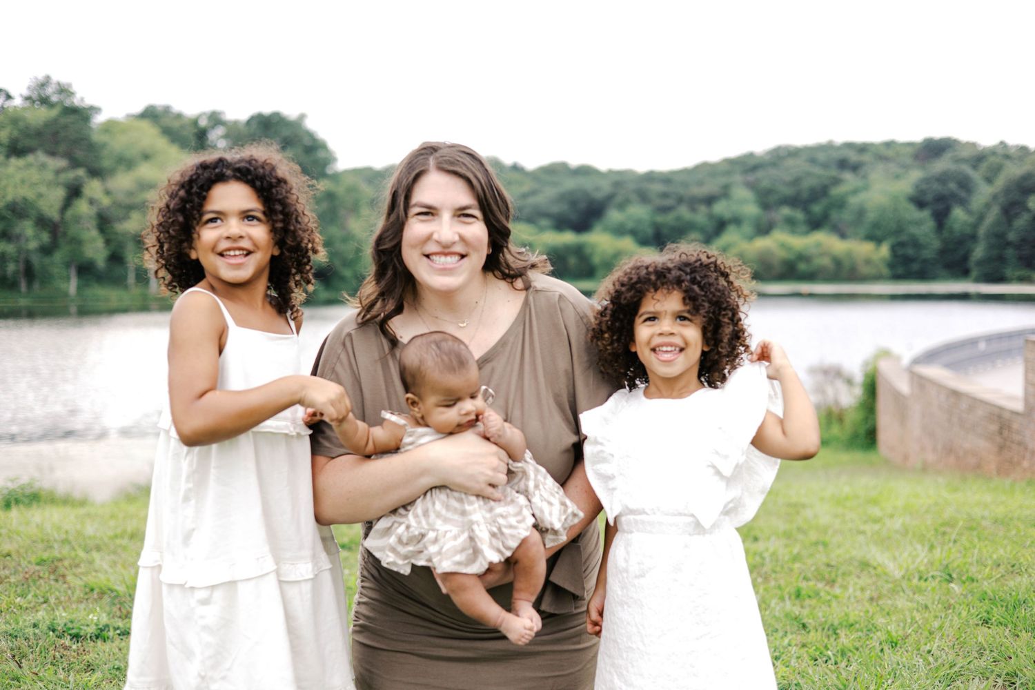 A woman holds a baby while two young girls with curly hair smile outdoors near a lake.