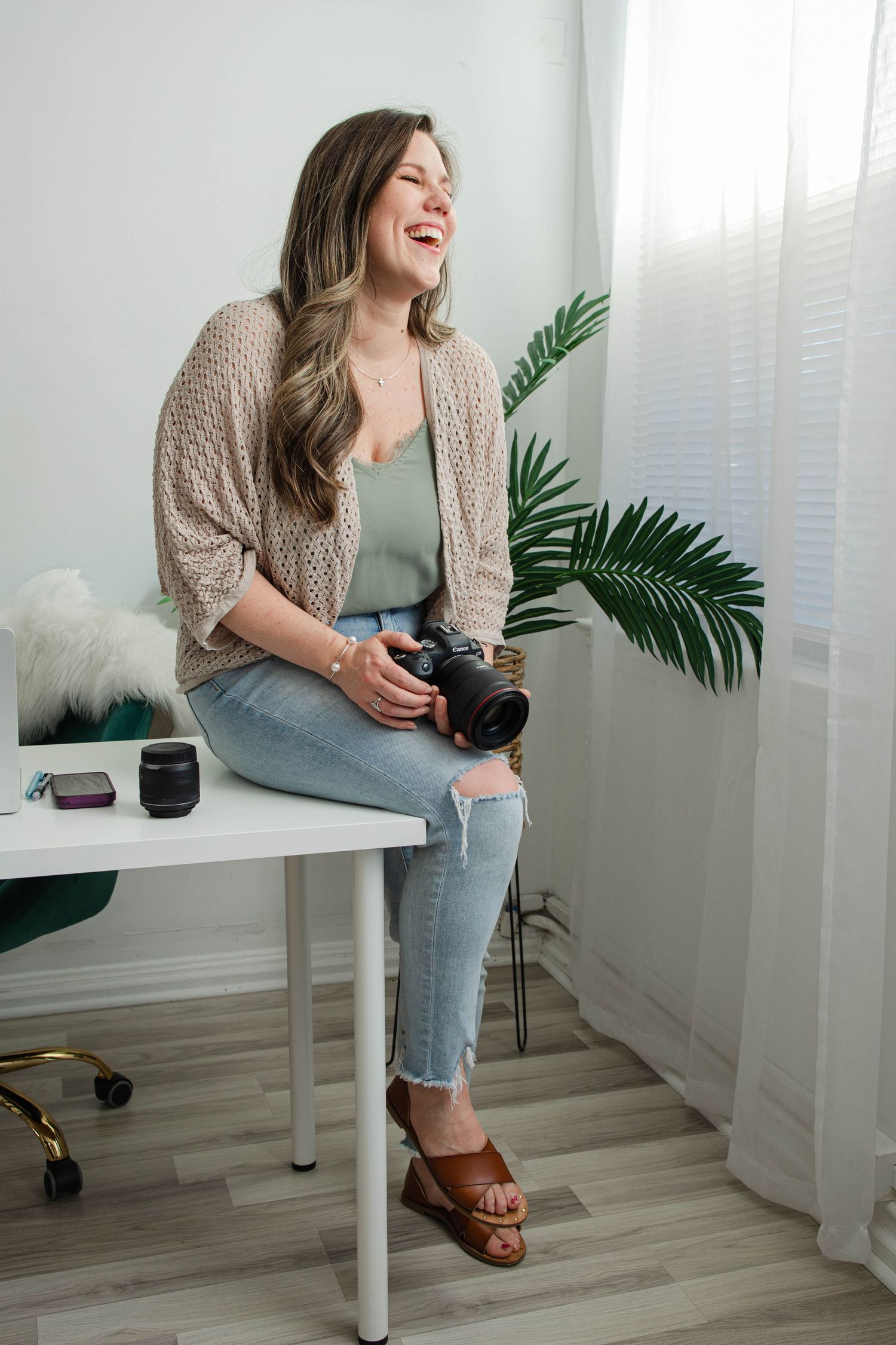 Photographer in beige cardigan poses with camera equipment on white desk with tropical plant.