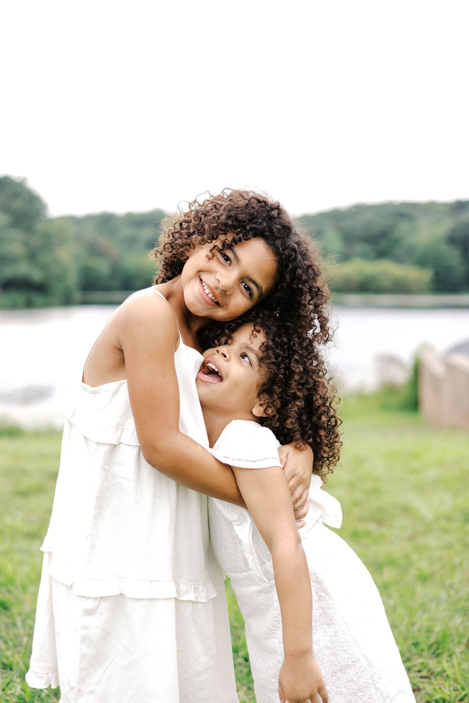 Two young girls with curly hair in white dresses hugging outdoors near a lake.