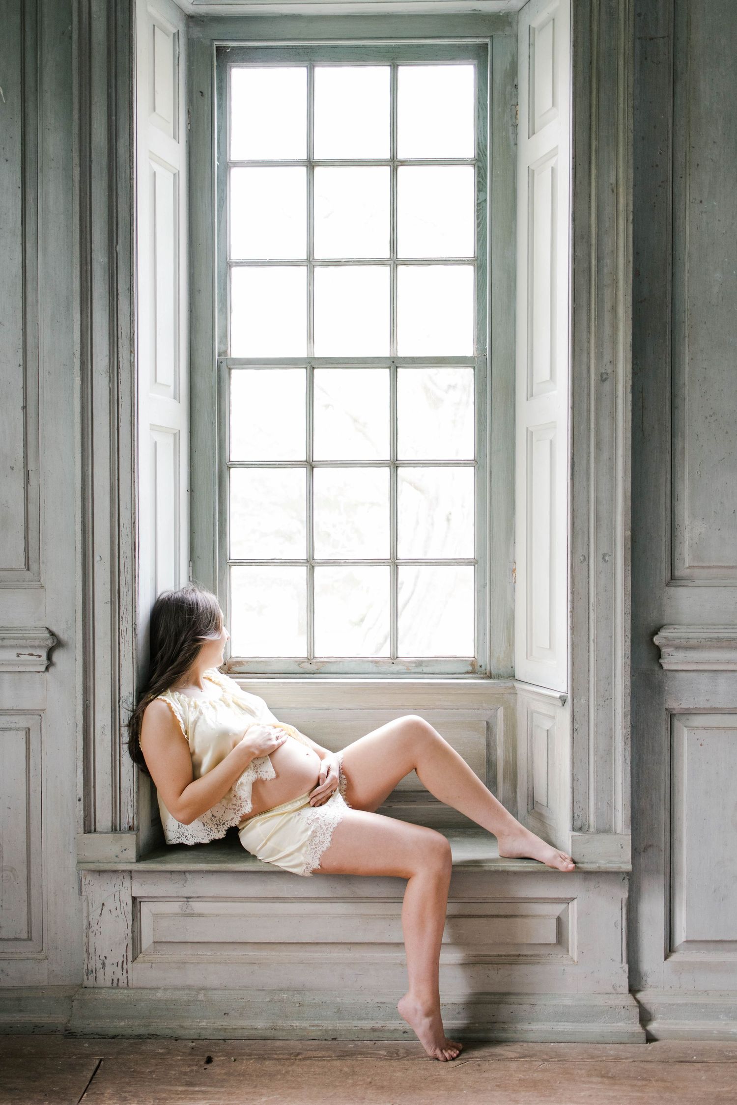 Pregnant woman in cream dress sitting barefoot on window seat in vintage room with tall white paneled windows.