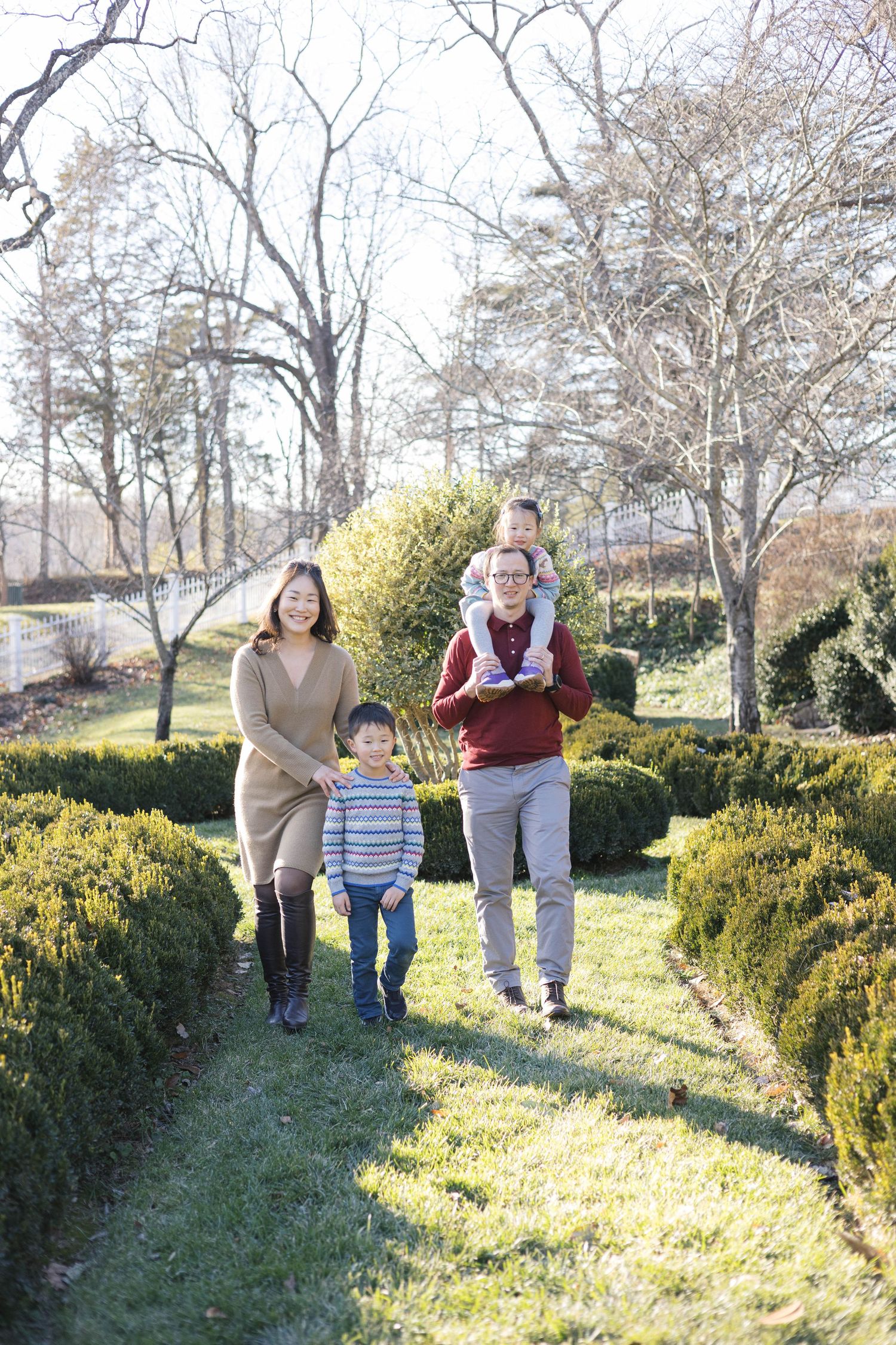 A family walks together on a garden path lined with manicured hedges on a sunny winter day.