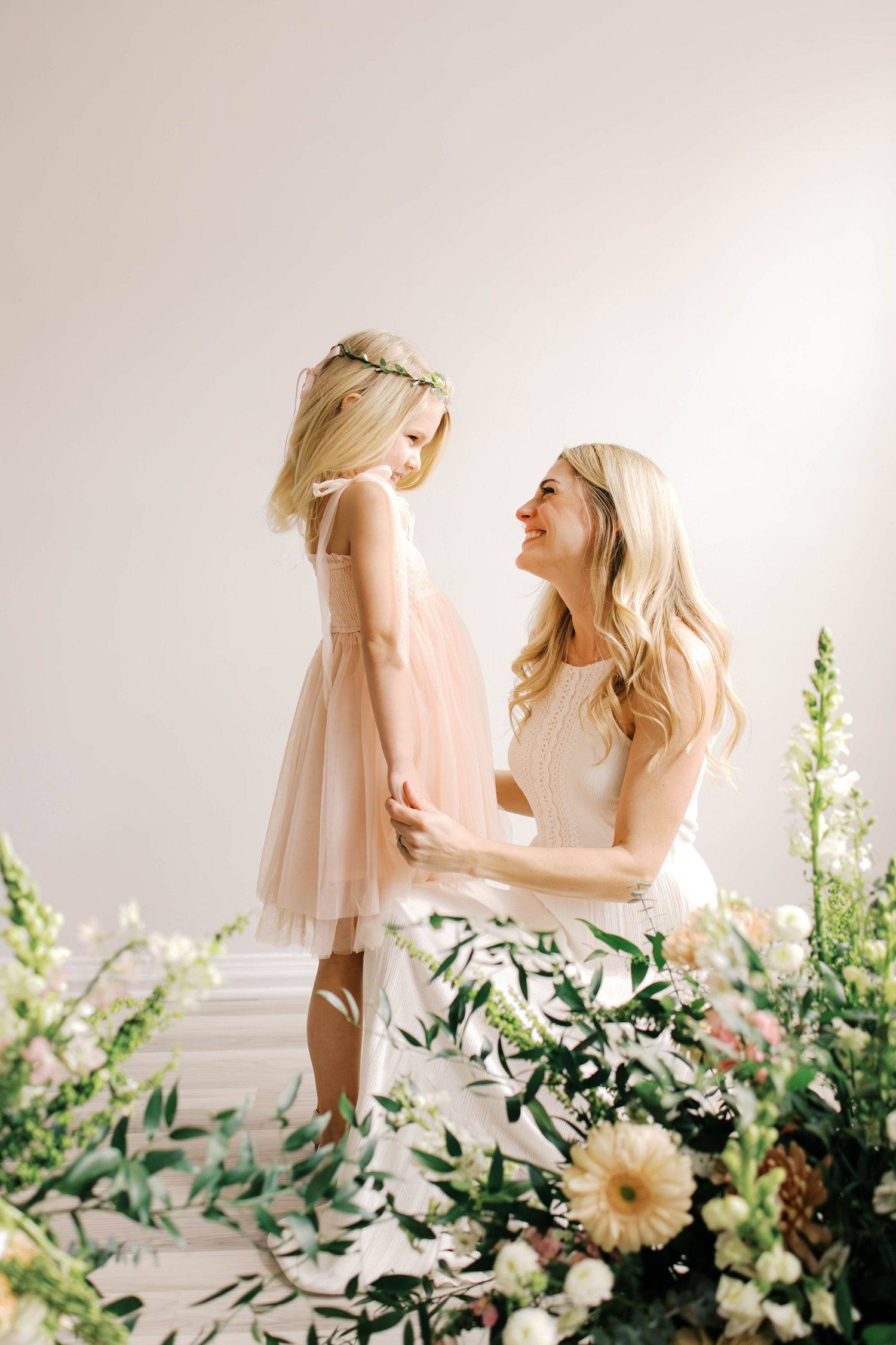 A smiling mother and her young daughter in a pink tulle dress and flower crown hold hands among white and cream blooms.