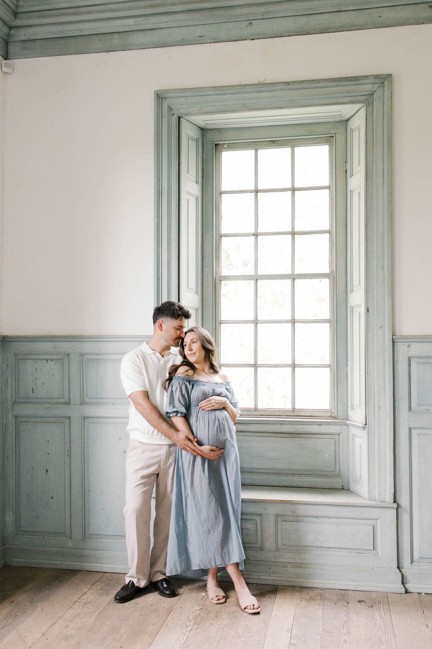 Couple sharing a tender moment by a vintage window, pregnant woman in blue dress cradling her bump in elegant room.