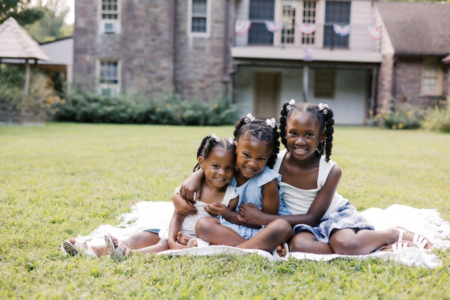 Three young girls sitting together on a blanket in a green yard, smiling and hugging in front of a brick house.