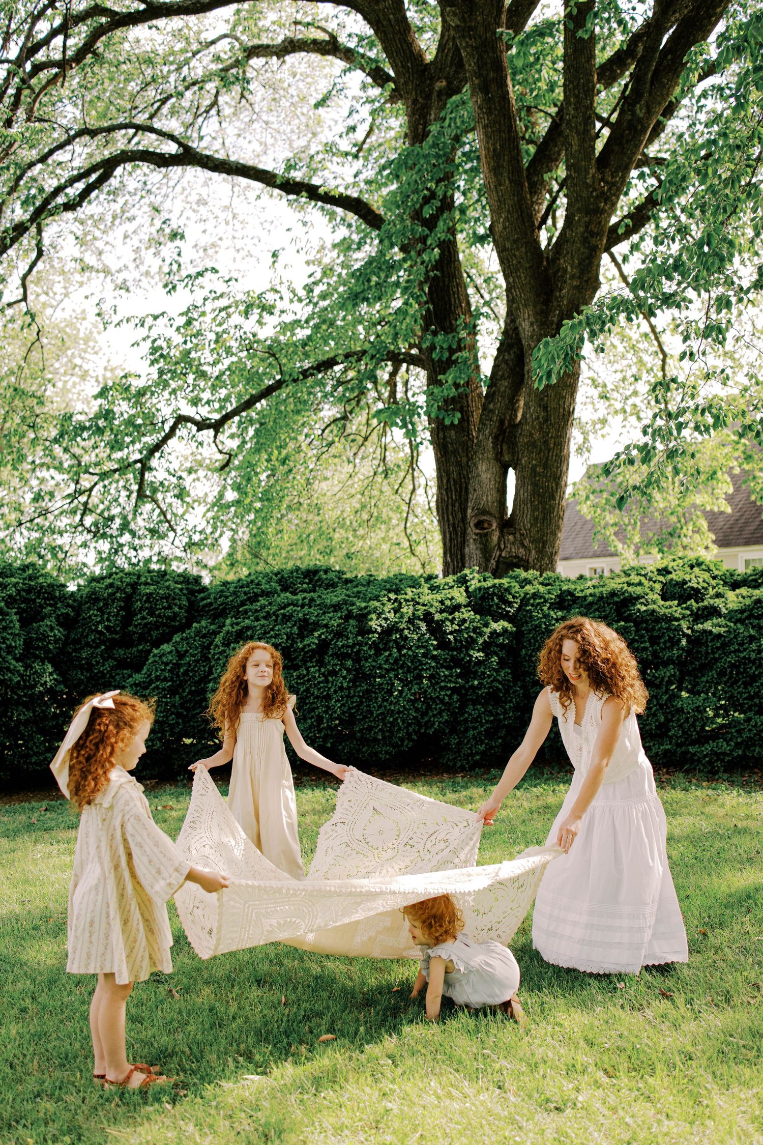 Four red-haired children in cream and white dresses spread a lace blanket on a sunlit lawn beneath a large tree.