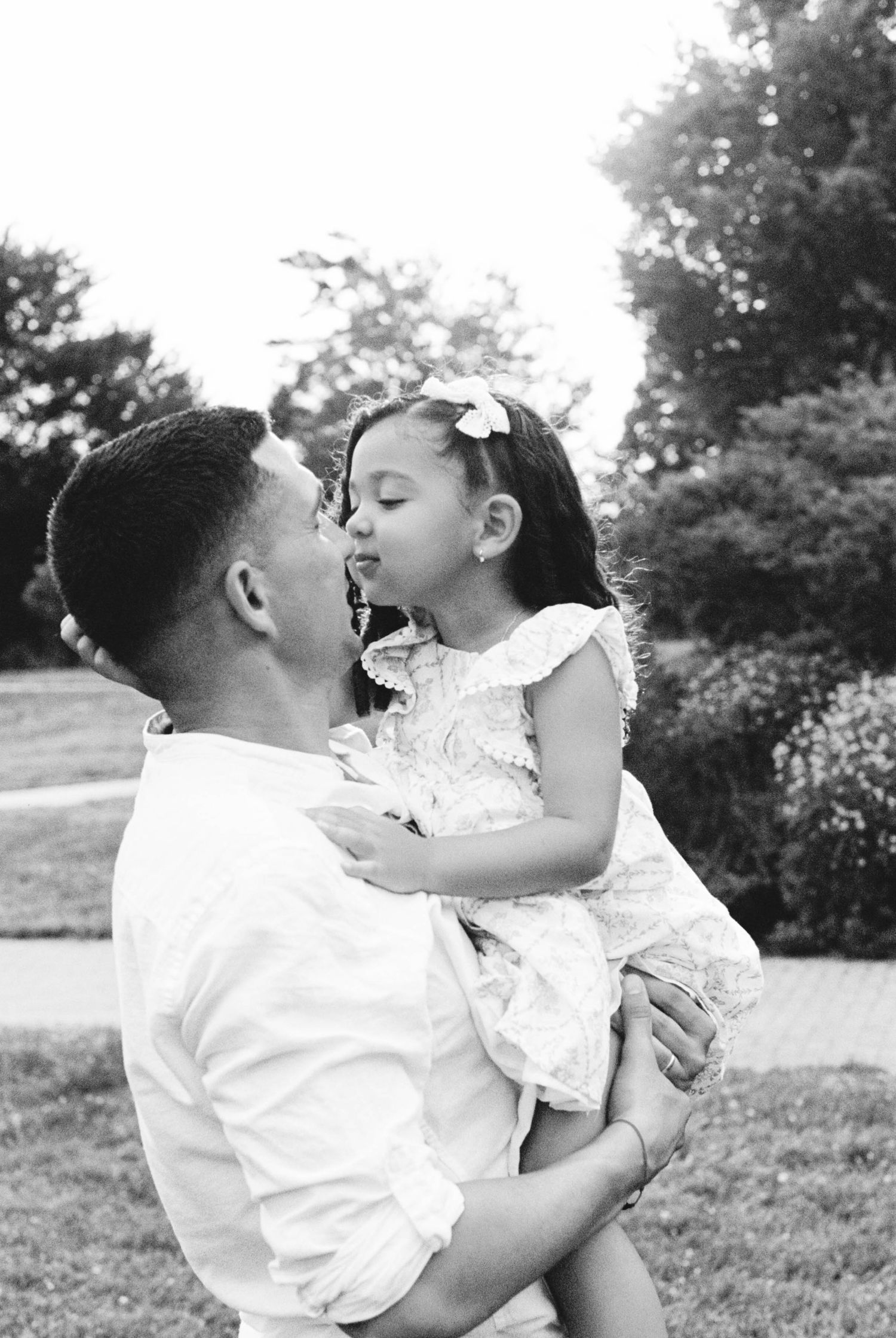 Black and white photo of a father holding his young daughter as she gives him a sweet kiss outdoors.