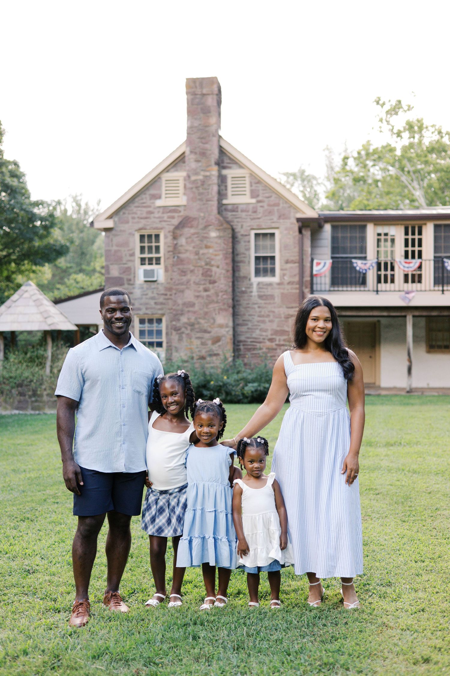 A joyful family of five poses outdoors on green grass in front of a brick home, dressed in coordinating light blue outfits.